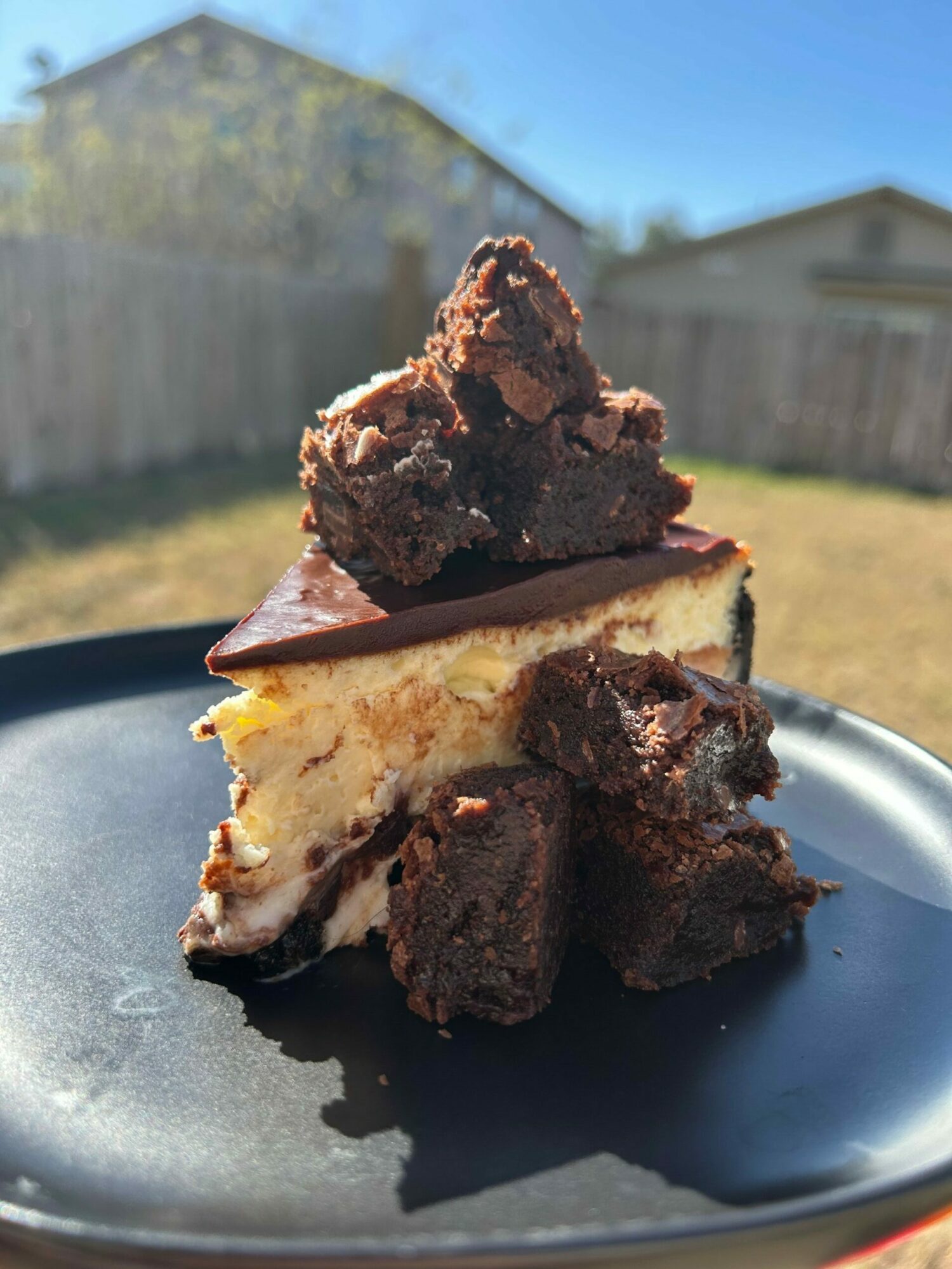 A slice of cheesecake topped with chocolate brownie pieces on a black plate outdoors.