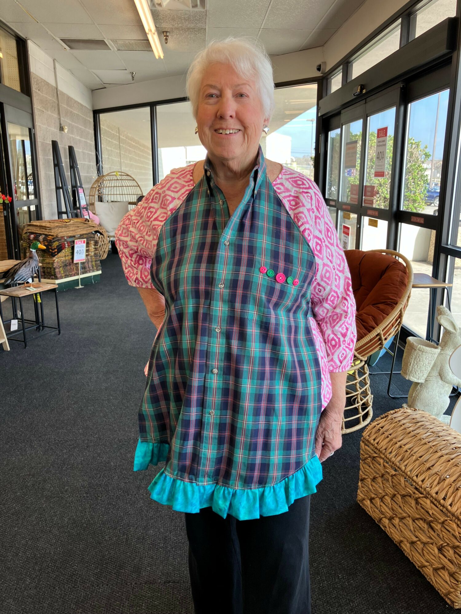 Older woman with white hair wearing a colorful plaid shirt and a pink patterned jacket, standing indoors near large windows.