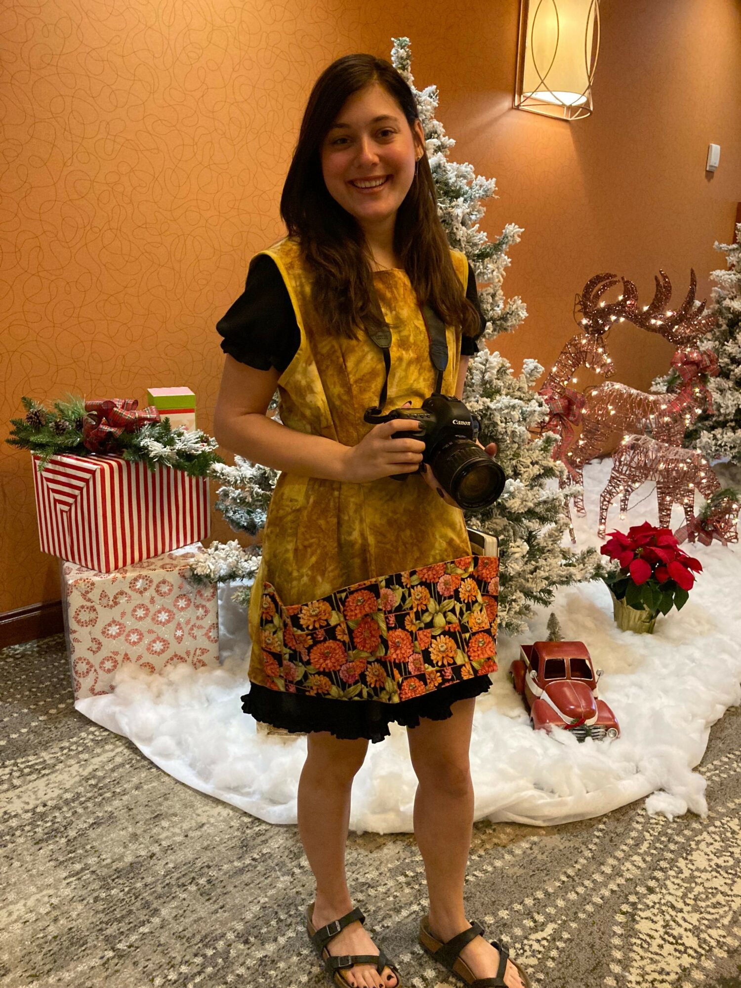 Young girl in a yellow dress with black sleeves and floral skirt, holding a camera, standing near a decorated Christmas tree.