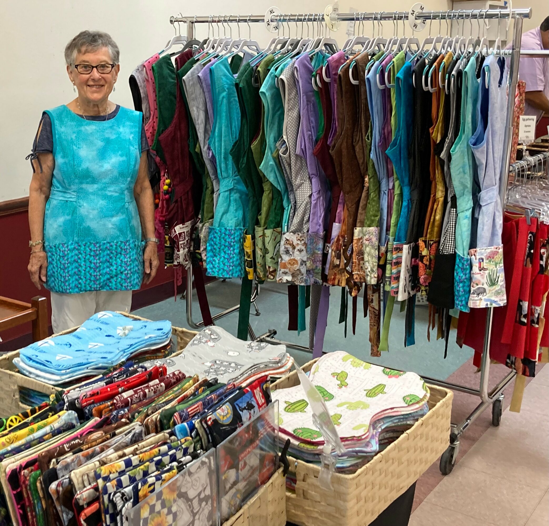 Elderly woman with short gray hair and glasses stands next to a clothing rack filled with colorful dresses, in a store.