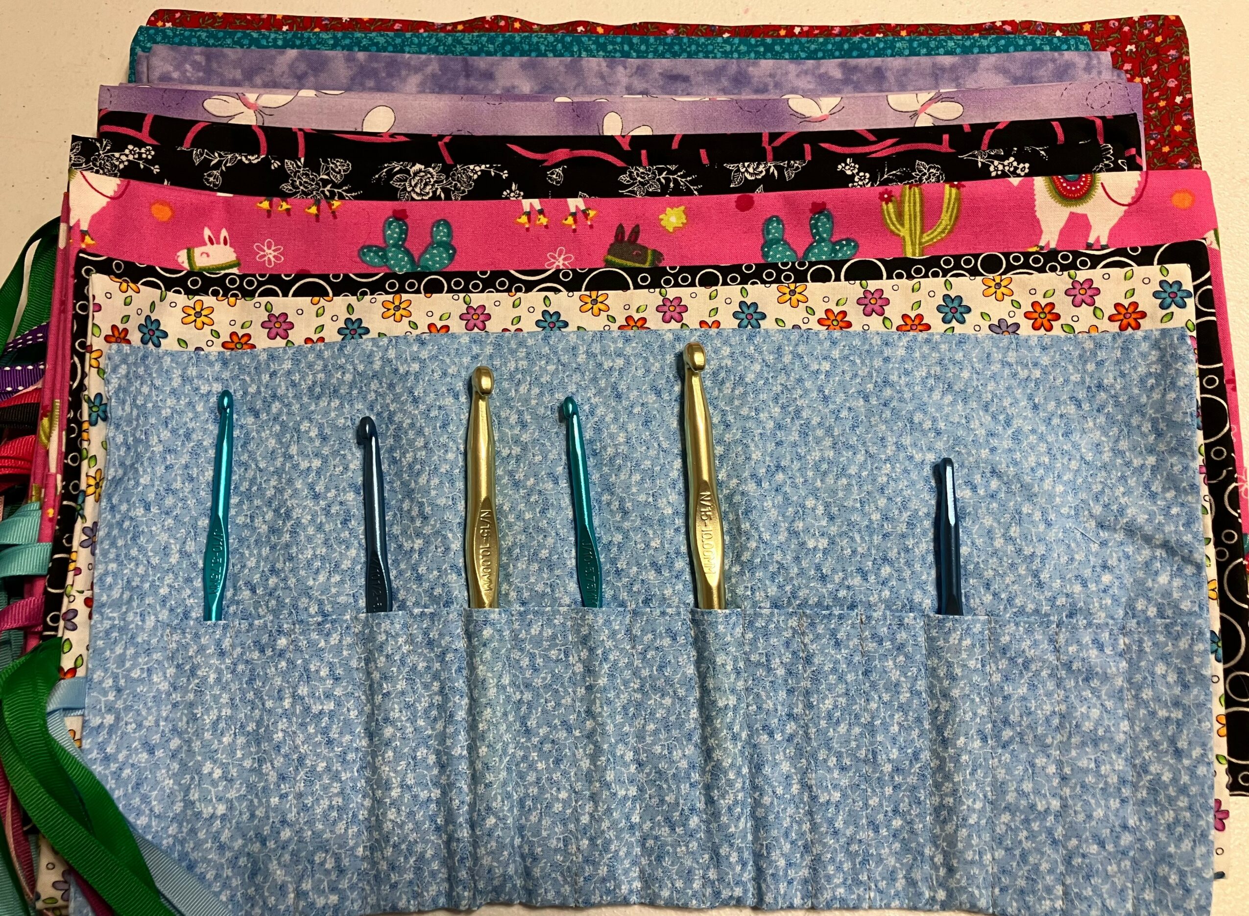 Multiple colorful fabric pieces stacked, with a blue fabric organizer holding five crochet hooks in front.