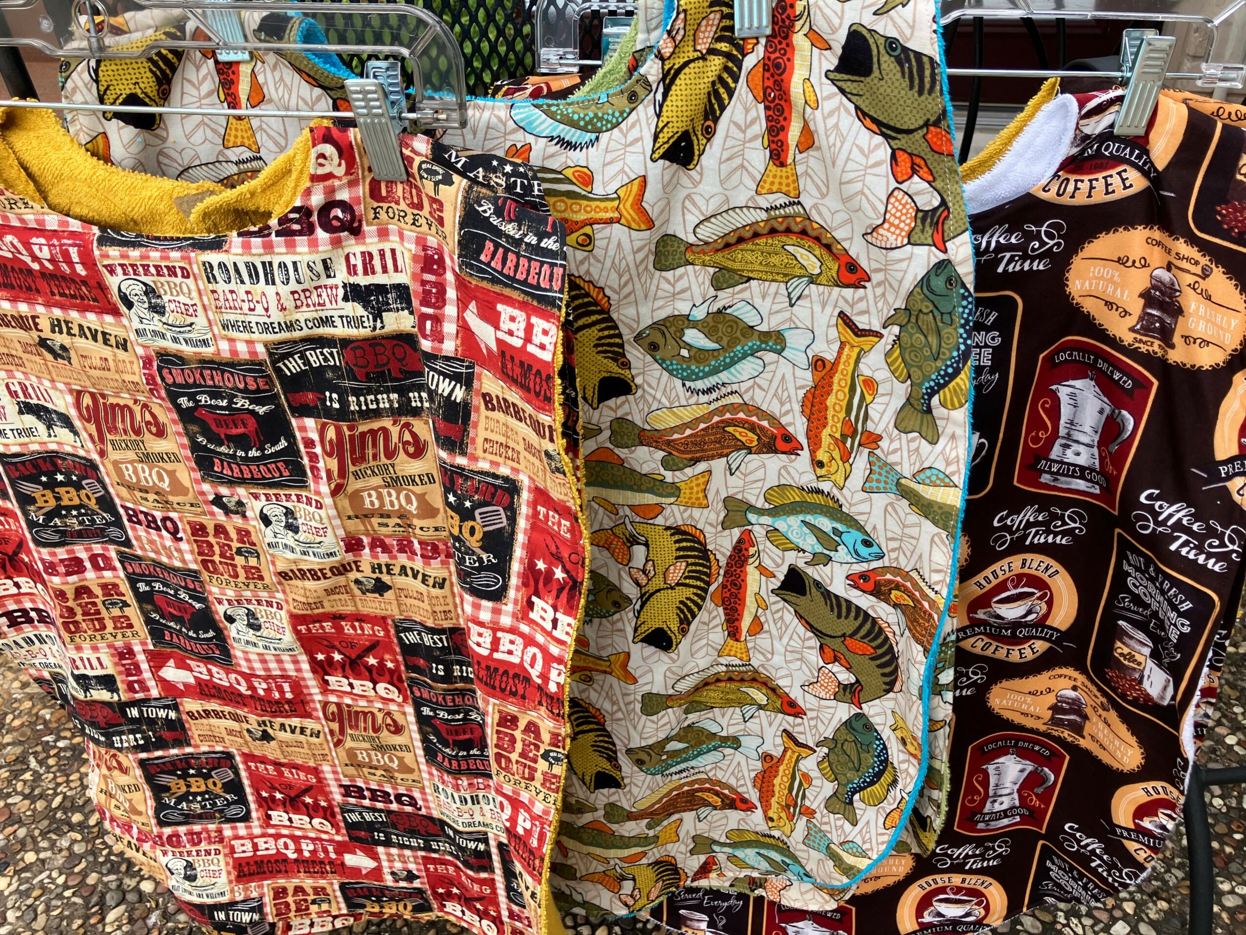 Three fabric bags with colorful patterns hanging on a rack, including a fish design, a barbecue theme, and a coffee motif.