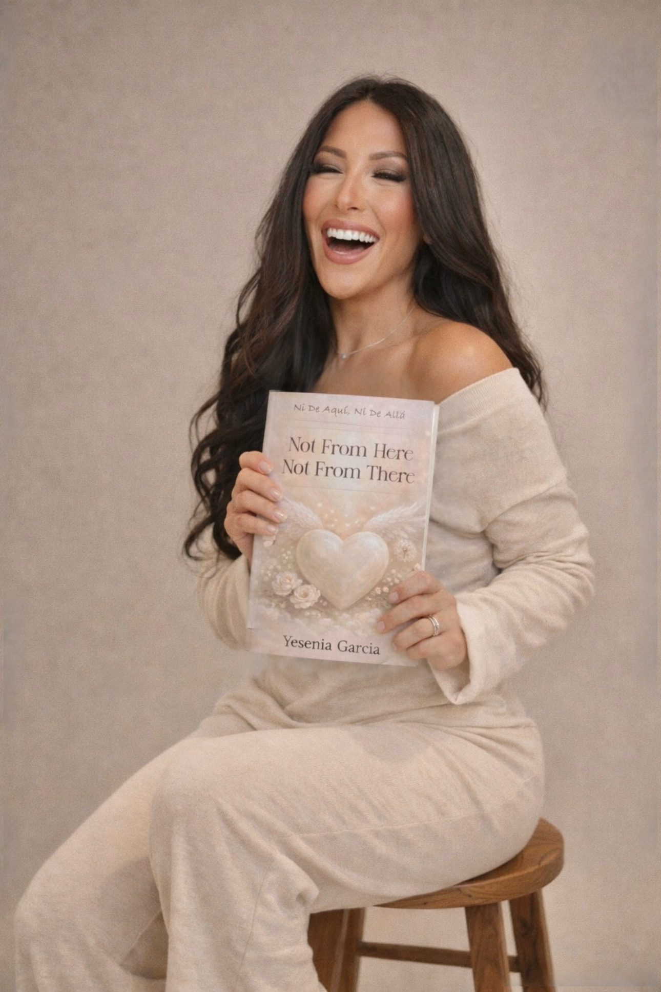 Woman with long dark hair smiling, holding a book titled 'Not From Here, Not From There,' sitting on a stool against a plain background.