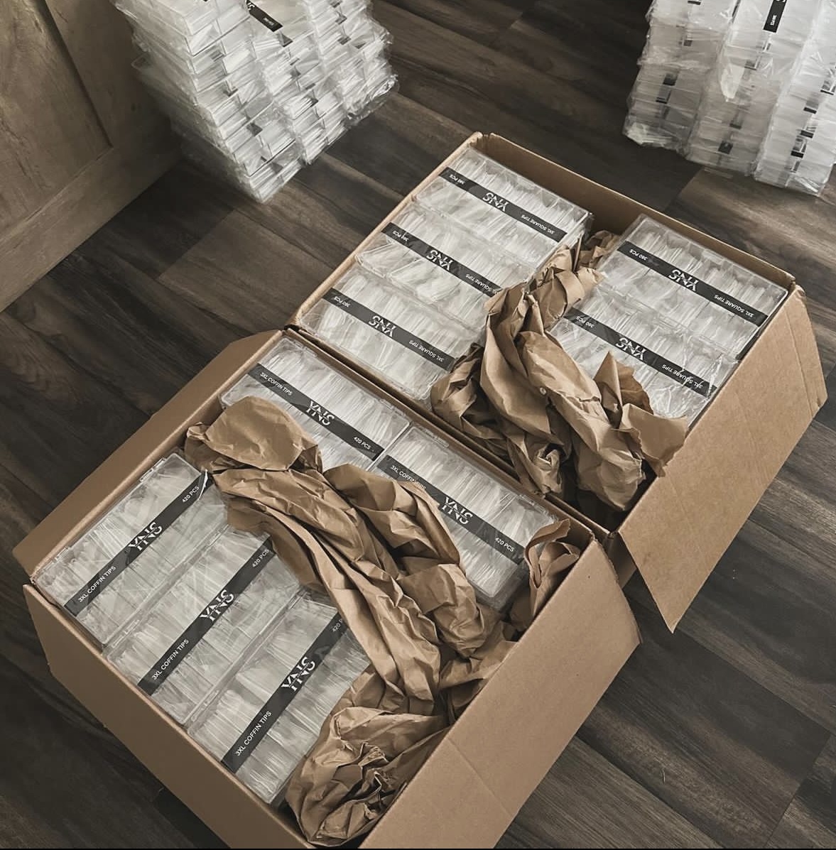Open cardboard box filled with neatly stacked packages wrapped in paper, on a wooden floor with more stacks in the background.