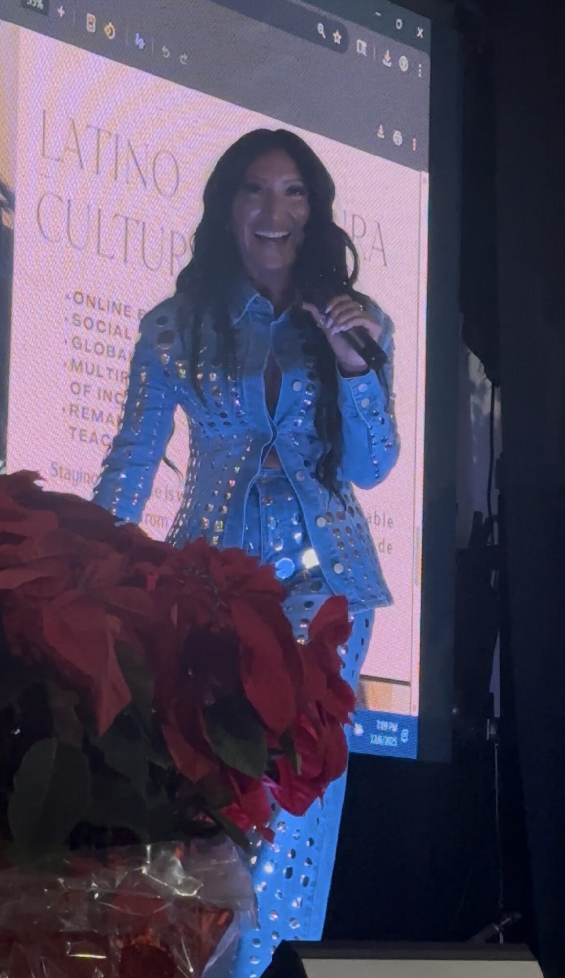 Woman smiling on stage with a microphone, wearing a blue outfit, behind a screen with text, and red flowers in foreground.