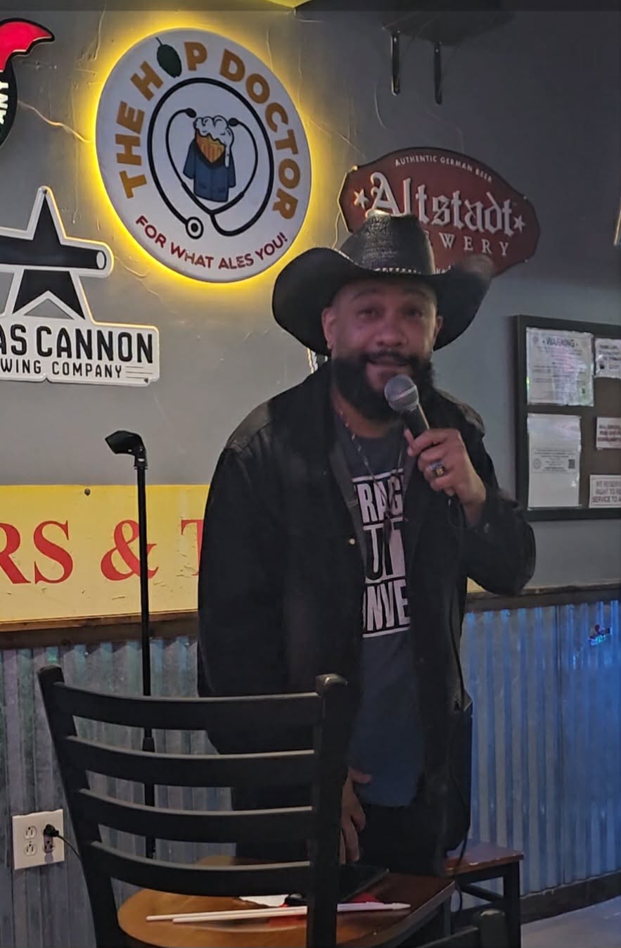 Man wearing a hat and jacket speaking into a microphone indoors, with signs and posters on the wall behind him.