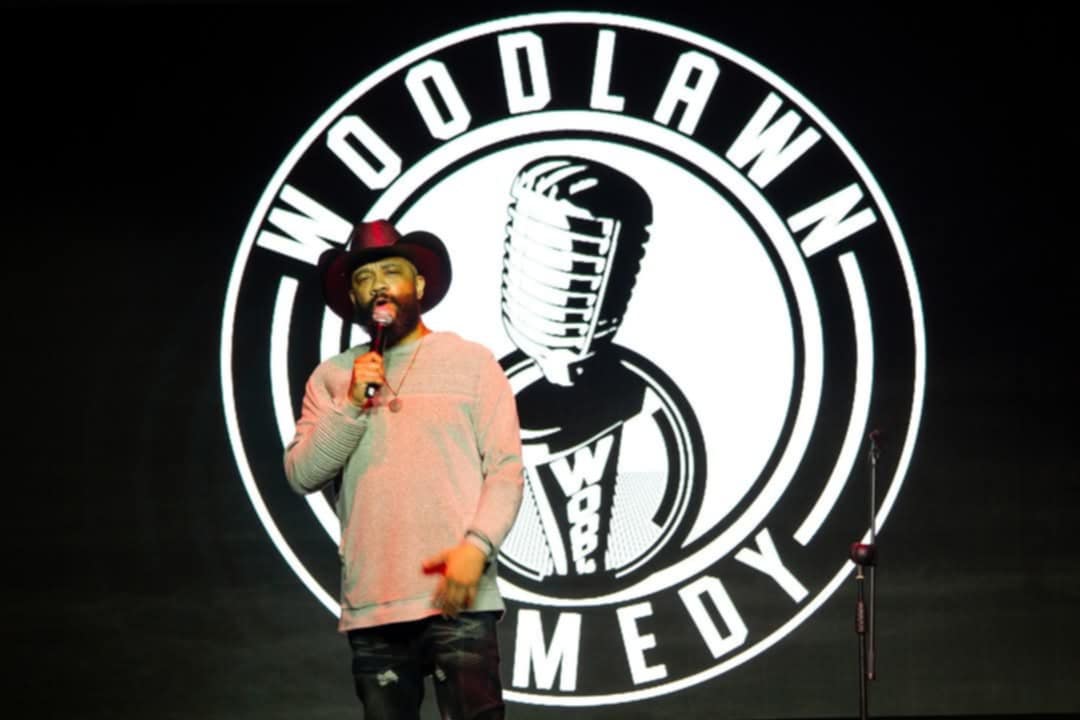 Person wearing a hat and sweater holding a microphone on stage with a circular logo behind them.
