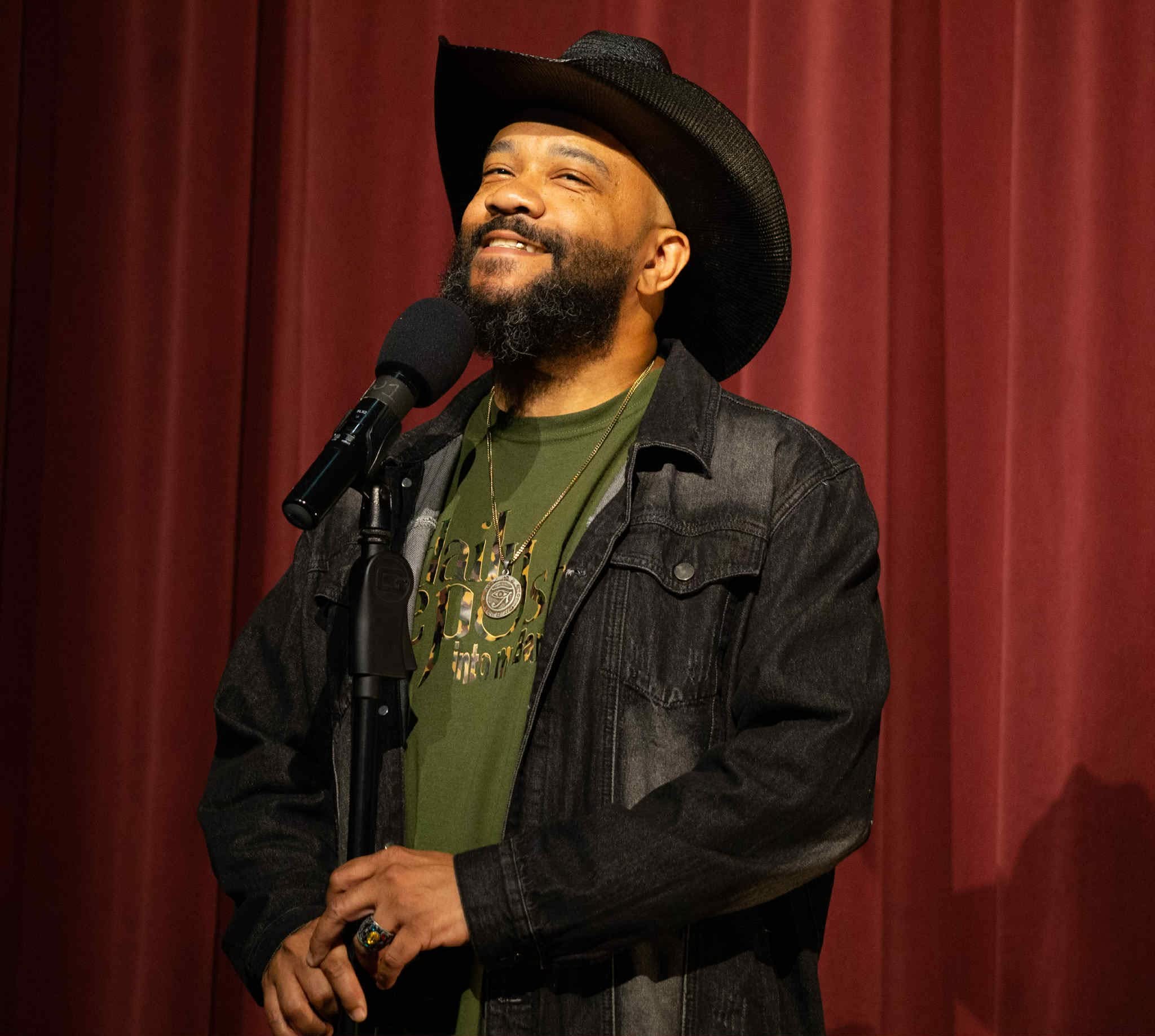 Man with a beard and mustache wearing a black hat, green shirt, and black jacket, speaking into a microphone on stage.