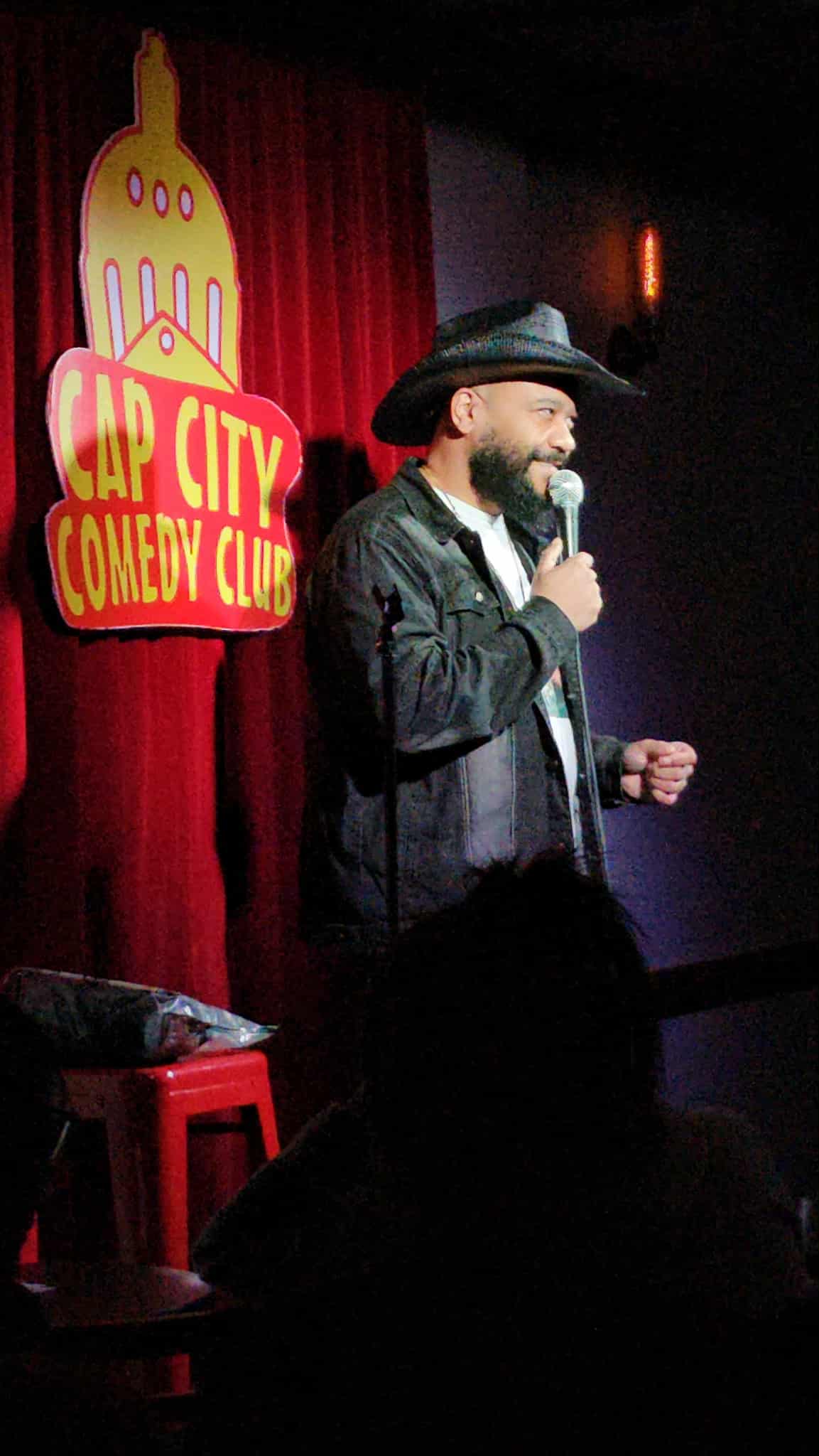 Comedian on stage holding a microphone, wearing a hat and jacket, with a red curtain and sign in the background.