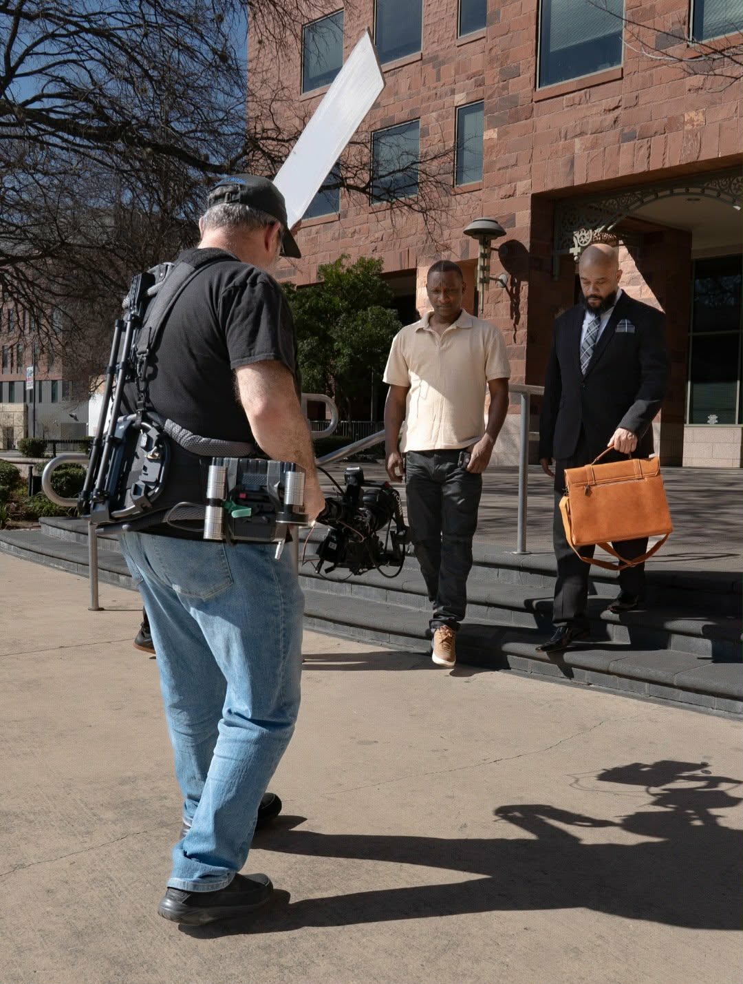 Photograph of three men walking outside a building, one carrying a briefcase, with shadows on the ground.