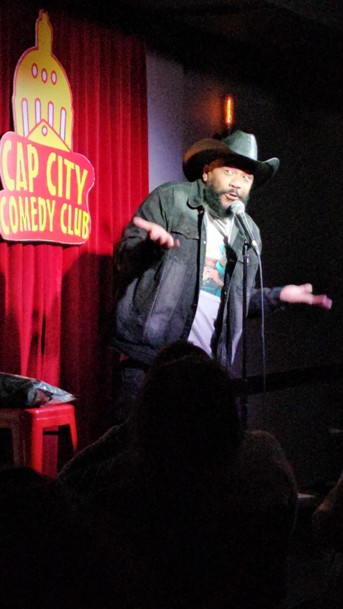 Comedian performing on stage at Cap City Comedy Club with red curtain background and logo, gesturing with hands.