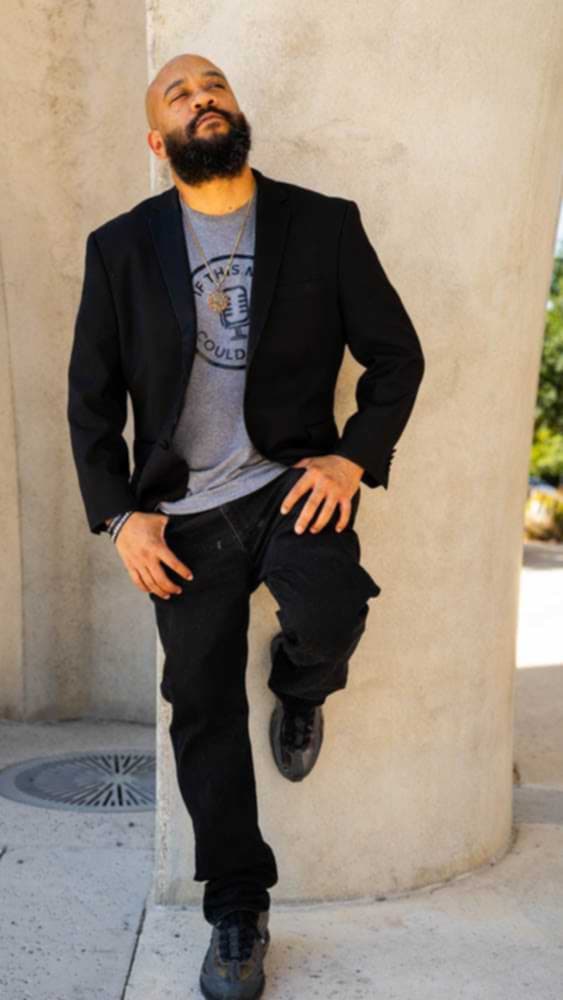 Man with beard and bald head leaning against a wall outdoors, wearing a black jacket, gray t-shirt, and black pants.