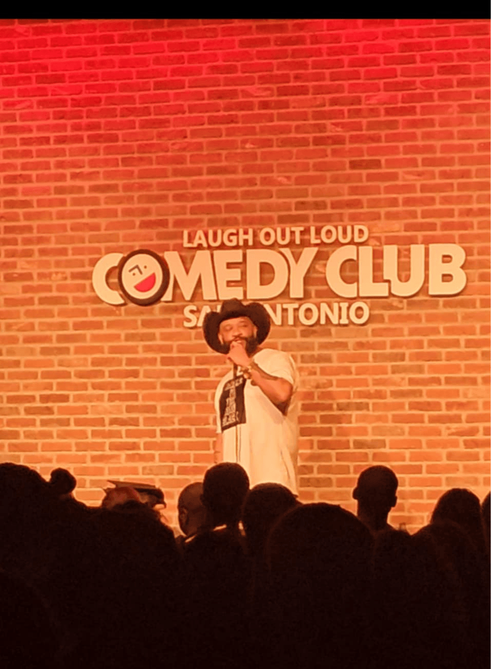 Stand-up comedian on stage with a brick wall background, audience silhouettes in foreground, comedy club sign behind.