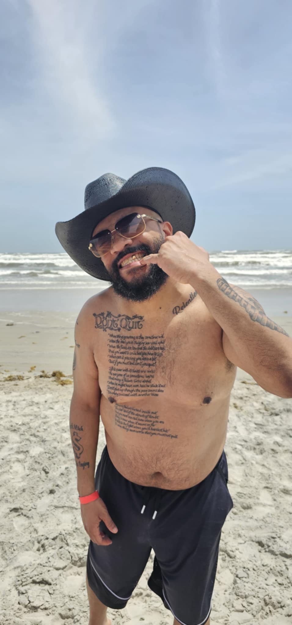 Man with a beard, sunglasses, and a wide-brimmed hat standing on a beach, smiling, with ocean waves in the background.