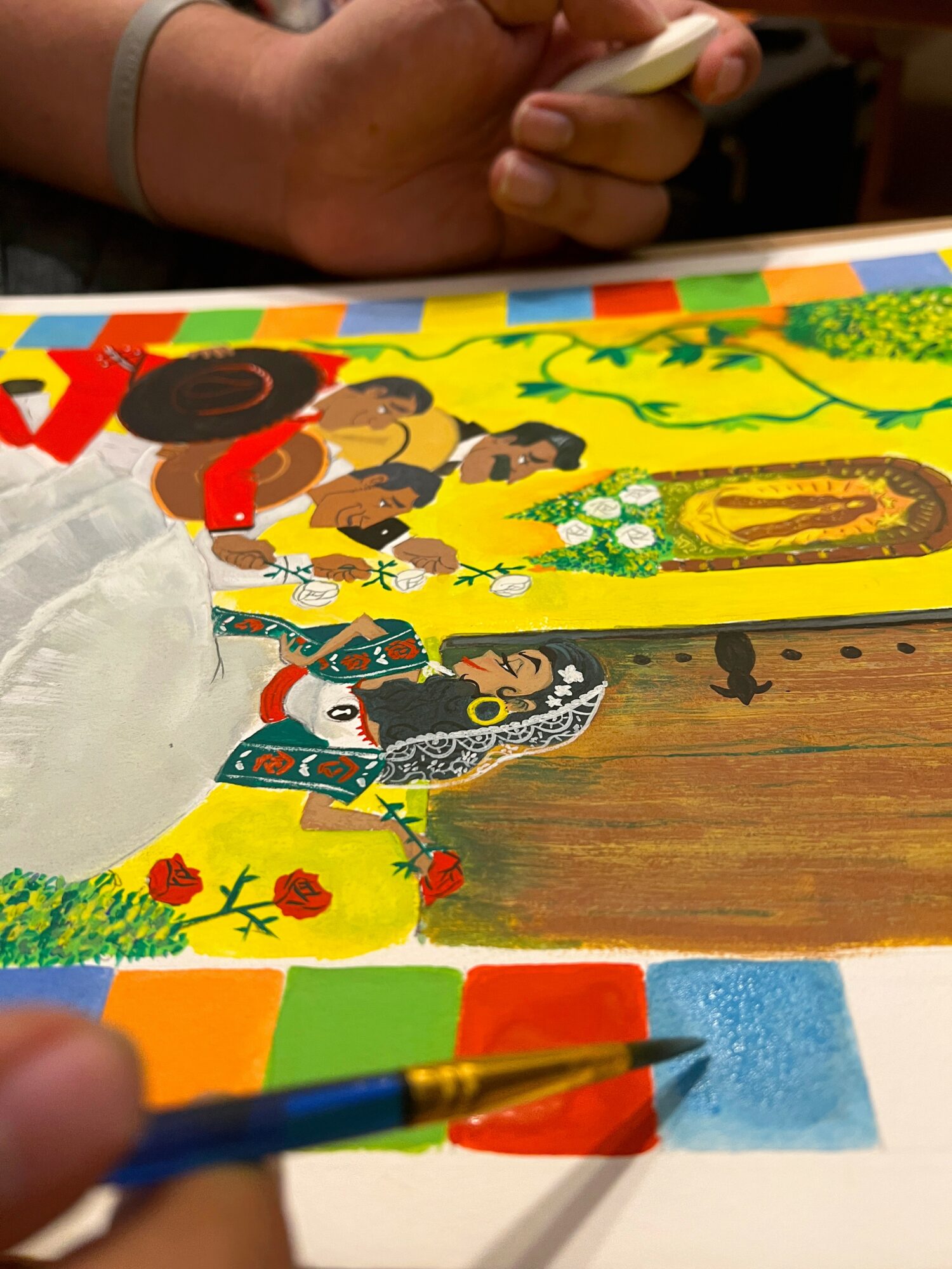 Colorful drawing on paper with various characters and scenes, placed on a wooden surface, with a person's hand holding a pen nearby.