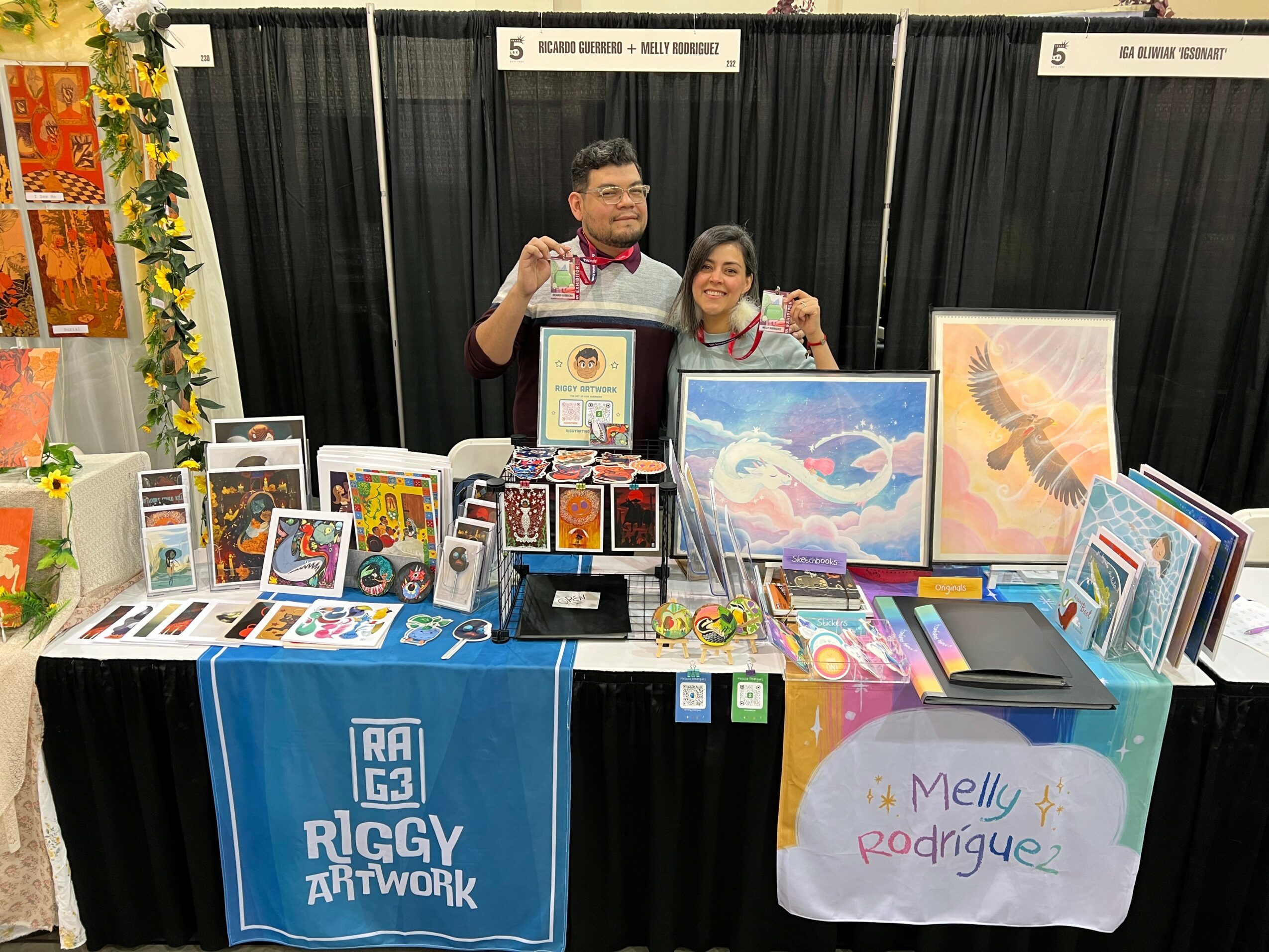 Two people stand behind a table with artwork and crafts at an event, black curtains in the background.