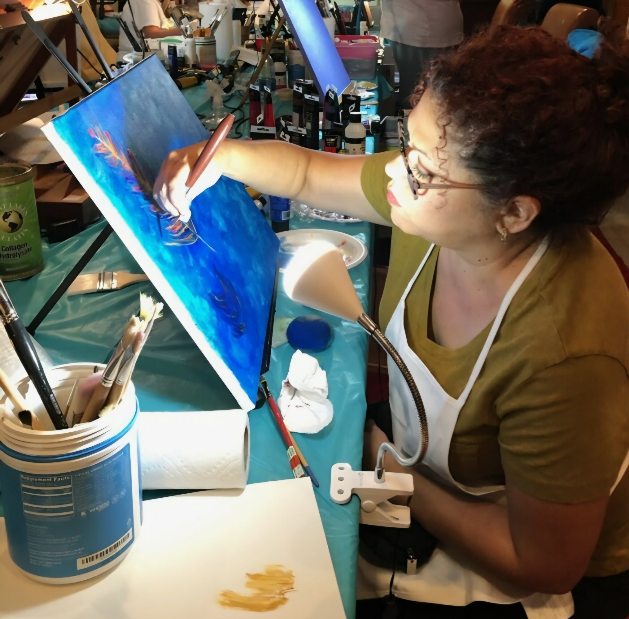 Woman painting on canvas at a cluttered art station, wearing glasses and a green shirt, with art supplies around her.