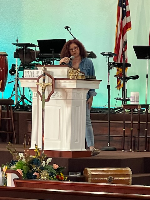 Woman standing behind a white pulpit on stage, with musical instruments and American flag in background, flowers in foreground.