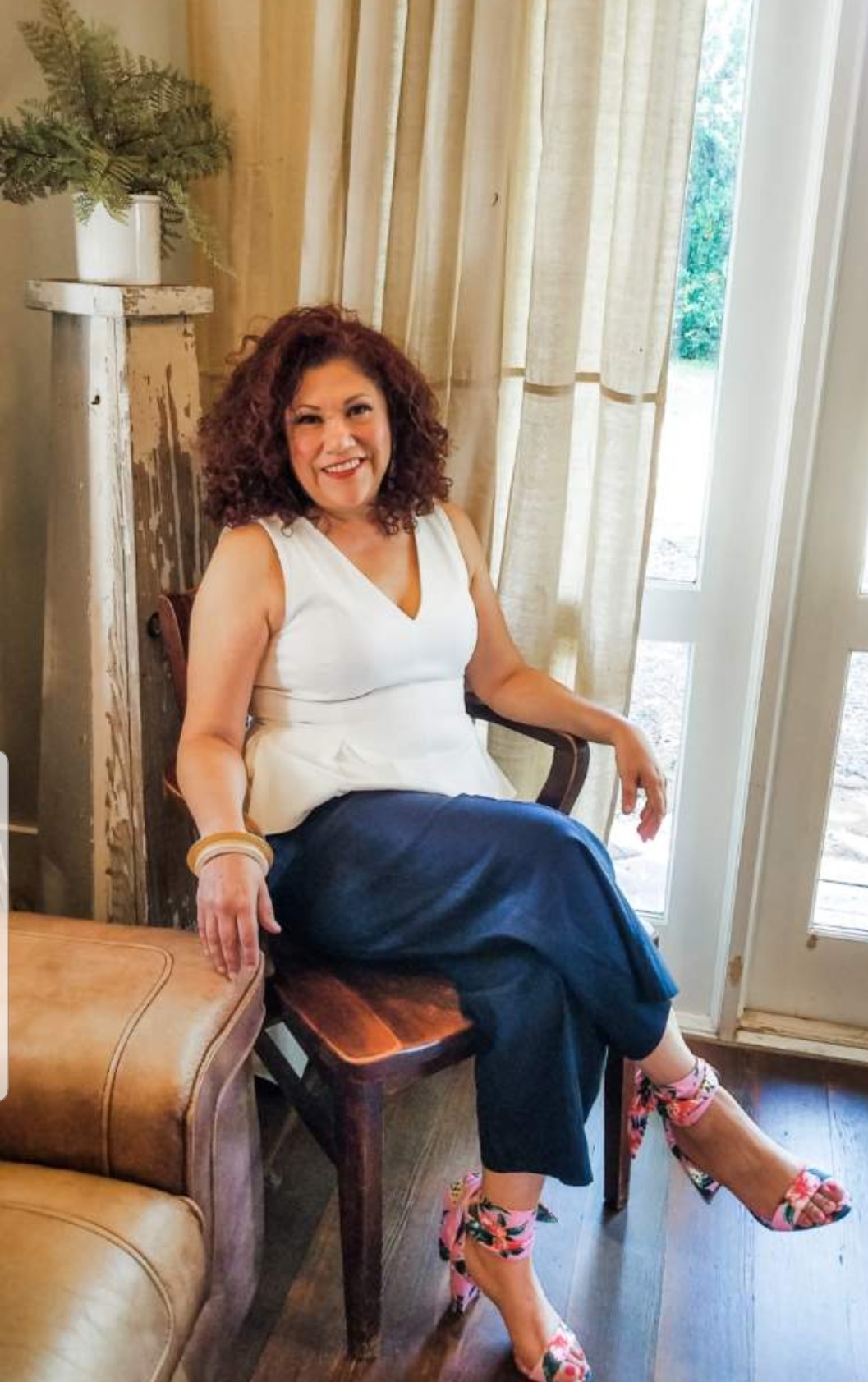 Woman with curly hair sitting on a wooden chair, smiling, in a room with curtains and a door, wearing a white top and dark pants.
