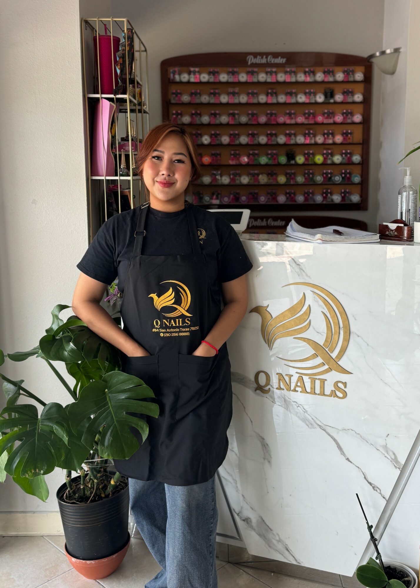 Woman standing behind a nail salon counter, wearing a black apron, with nail polish display behind her, and a potted plant nearby.