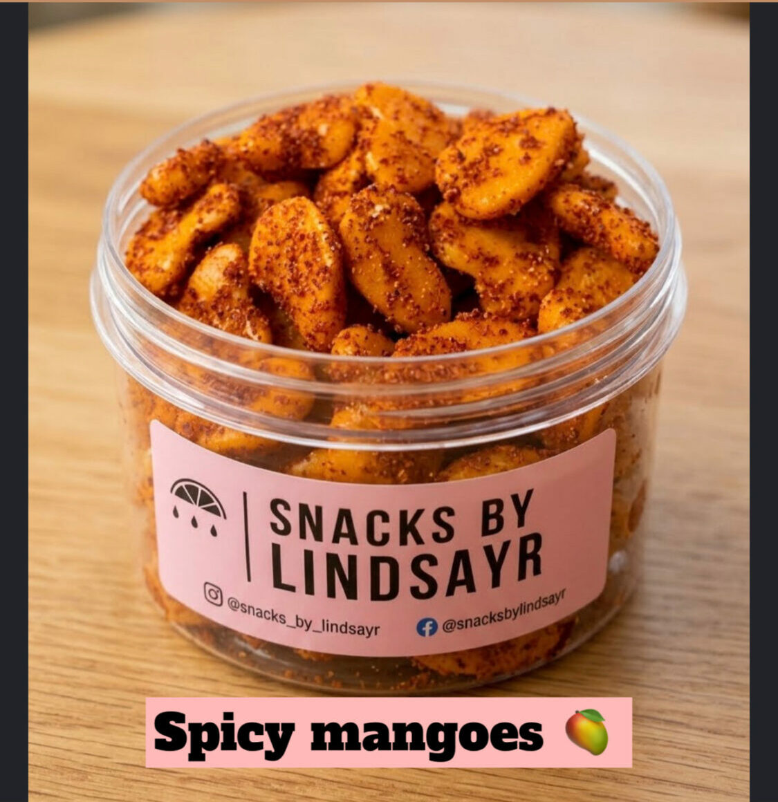 Container of spicy mango-flavored snacks on a wooden surface, label reads 'Snacks by Lindsay R,' with a pink banner 'Spicy mangoes' and a mango icon.