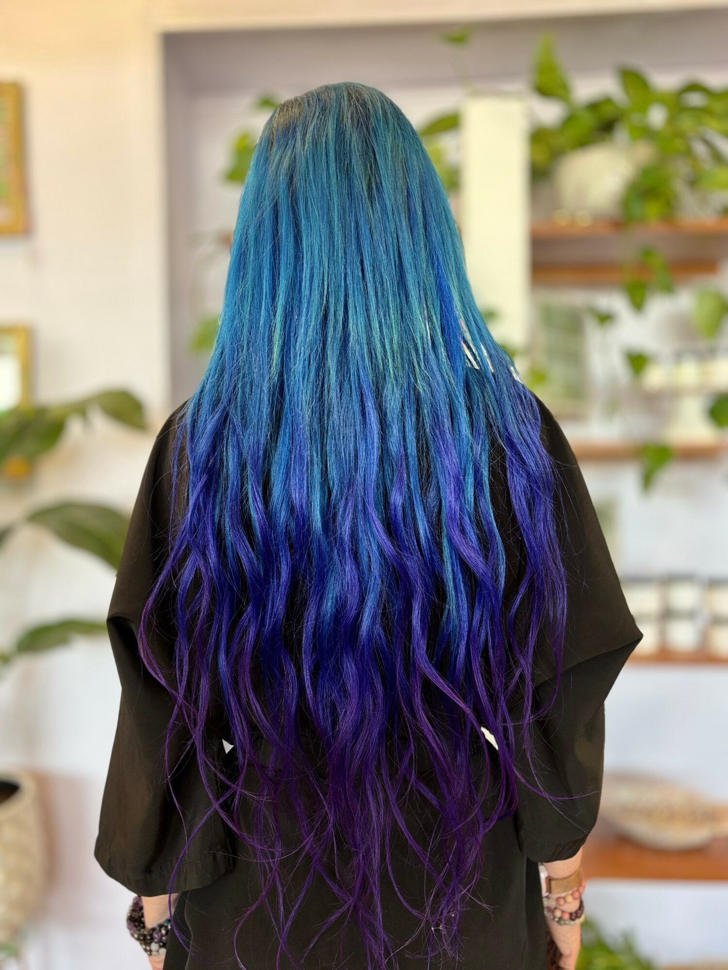 Person with long, wavy hair dyed in shades of blue and purple, standing indoors with plants and shelves in background.