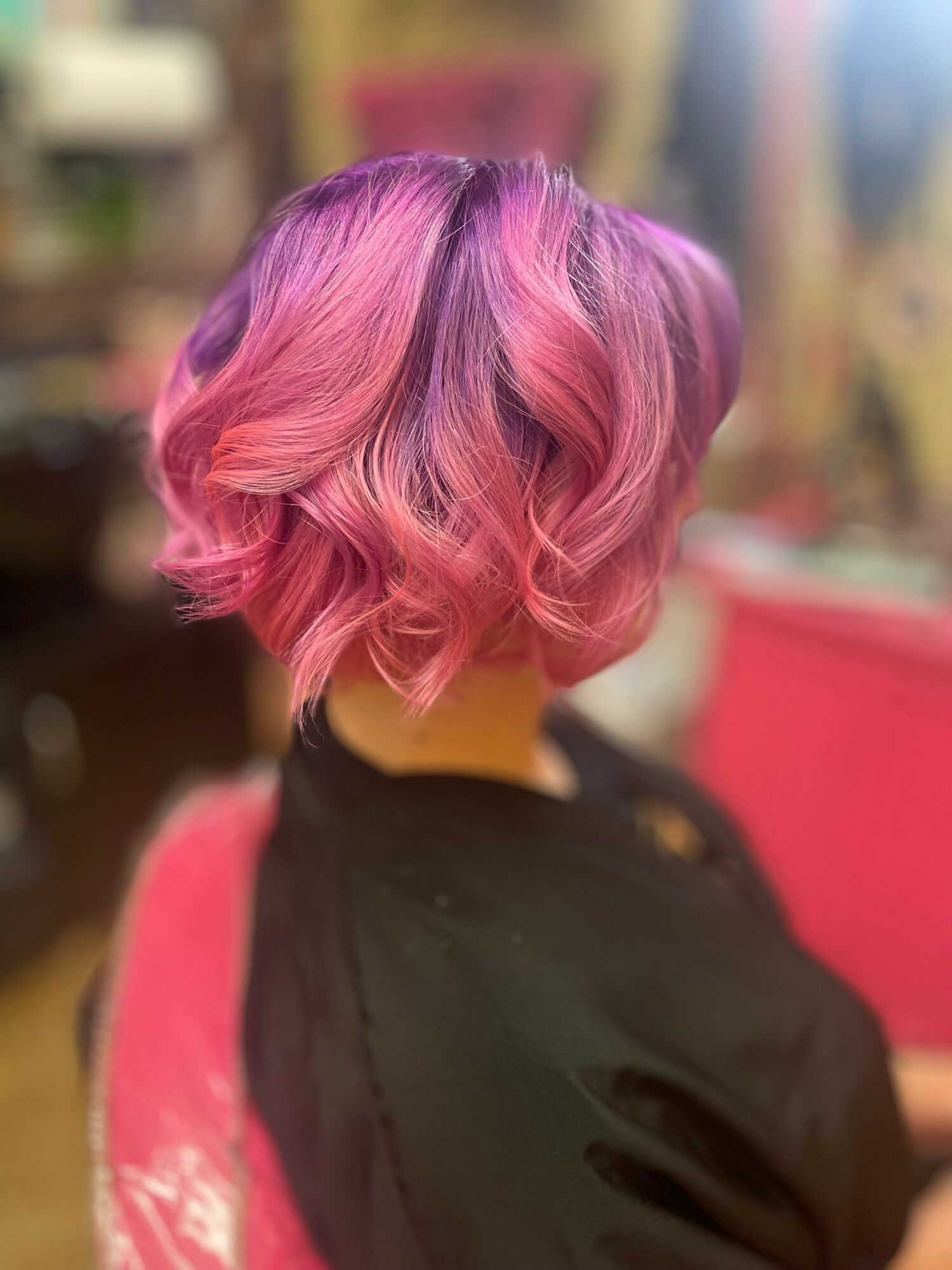 Back view of a person with pink and purple wavy, shoulder-length hair, wearing a black top.