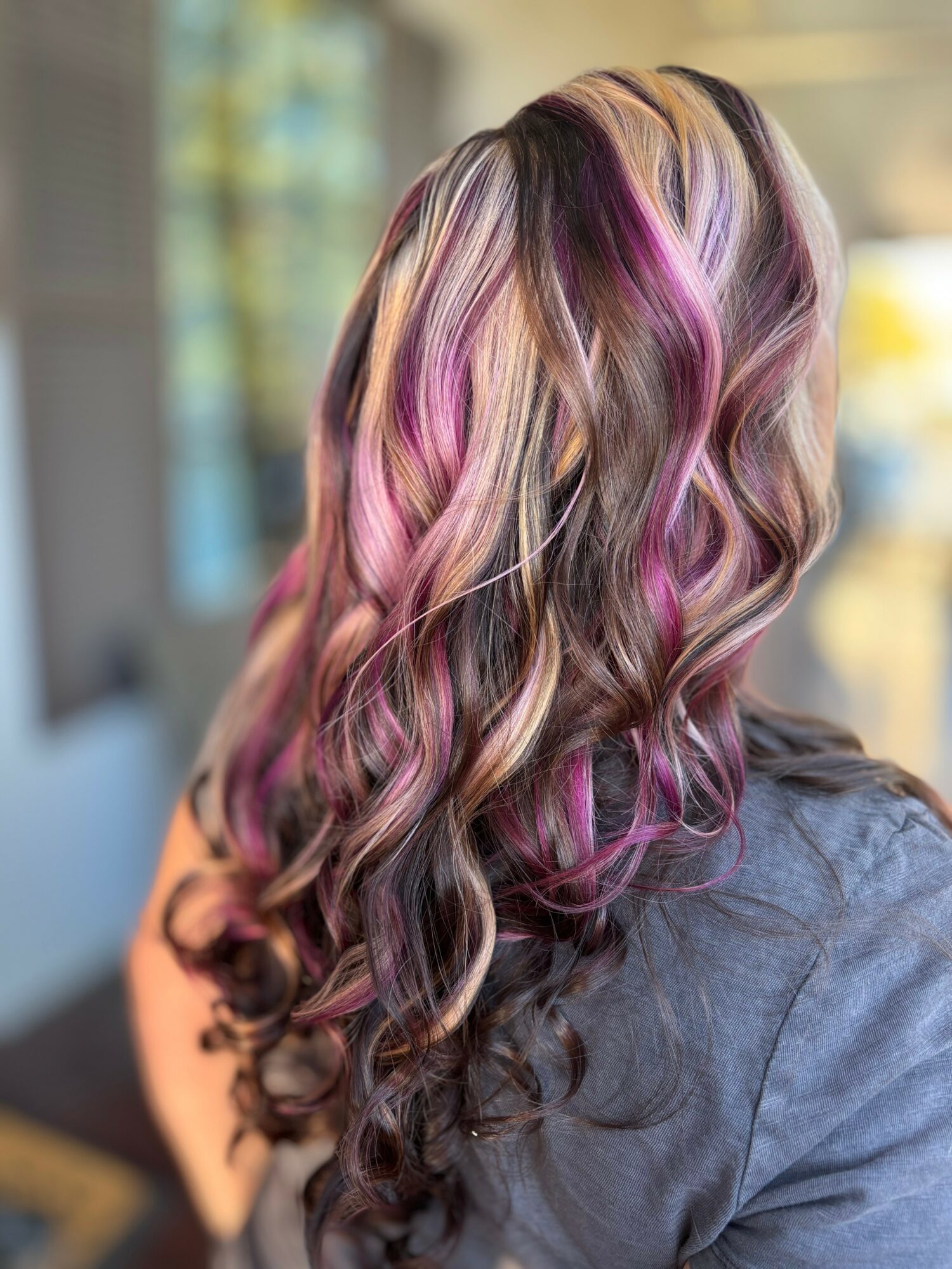 Person with long, wavy hair featuring blonde, pink, and purple highlights, viewed from the back.
