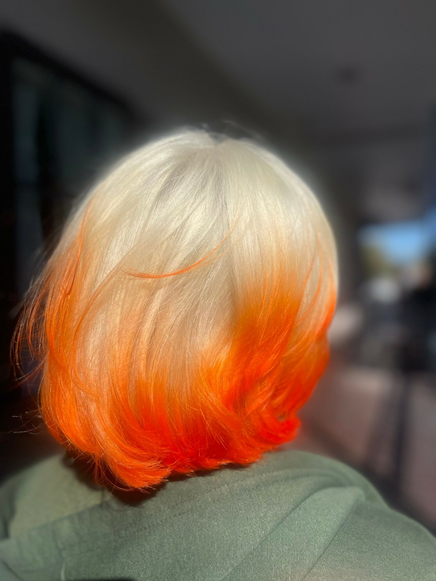 Back of a person's head with blonde hair fading to orange at the tips, indoors with blurred background.