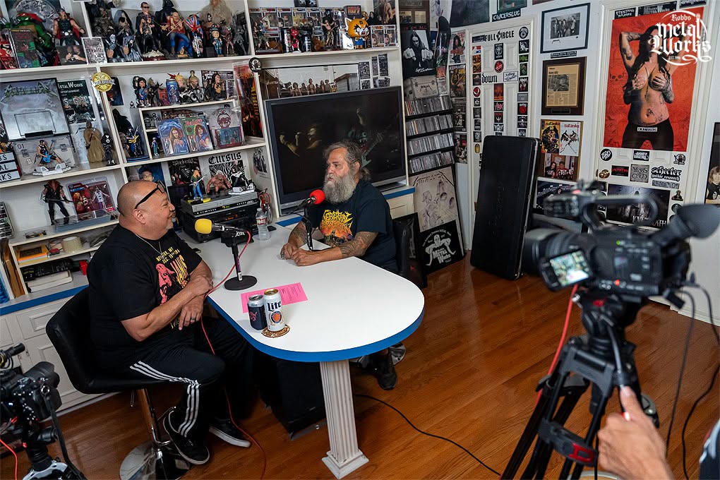 Two men sit at a round table in a room filled with shelves of collectibles and posters, recording a conversation.