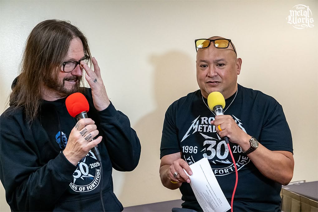 Two men speaking into microphones, one with glasses and long hair, the other bald with sunglasses on head, holding papers.