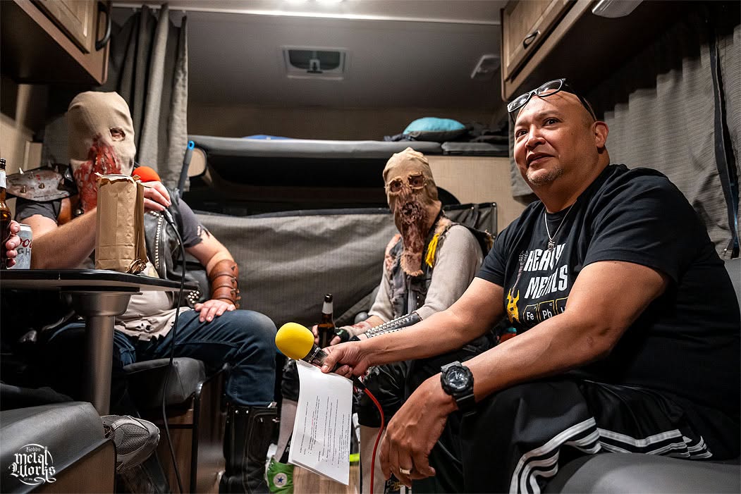 Three people sitting inside a vehicle, two women and one man, engaged in conversation, with a bed and curtains in the background.