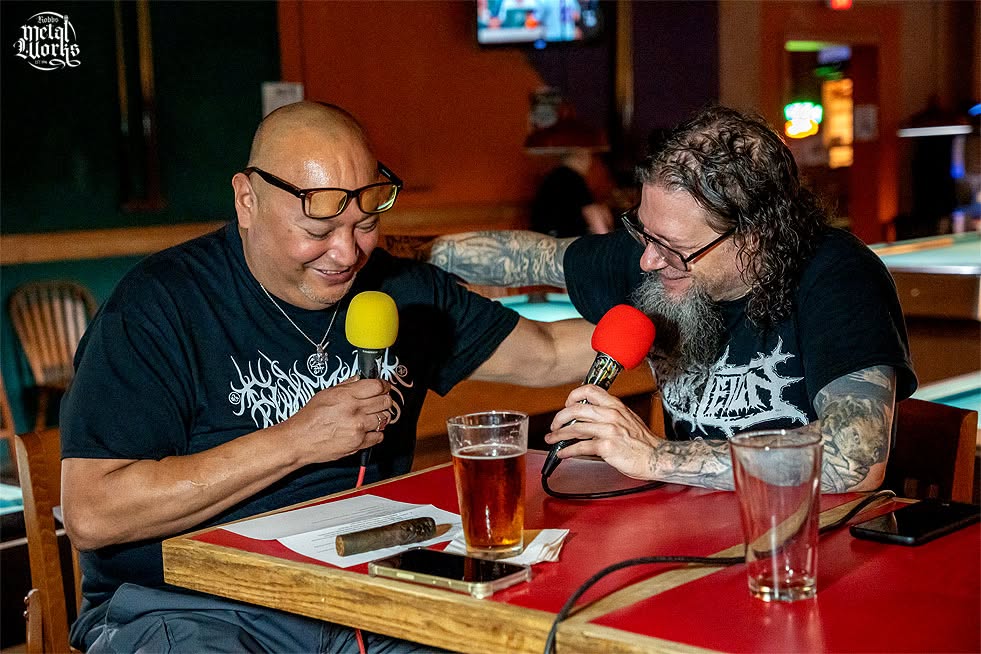 Two men with glasses and tattoos sit at a bar, holding microphones with yellow and red foam covers, talking and smiling.