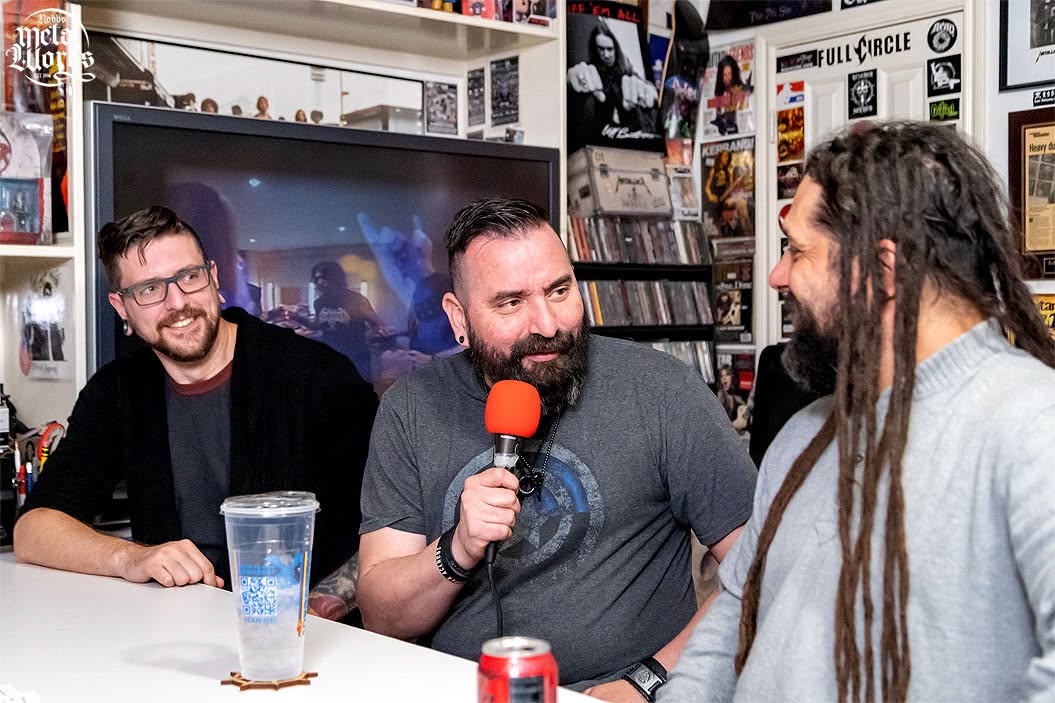 Three men sit at a table, one holding a microphone with a red cover, in a room with posters and a TV screen.