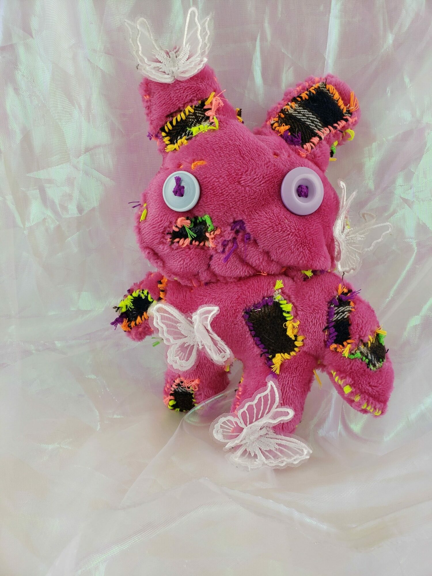 Pink plush toy with button eyes, colorful striped ears, and white wings, lying on a shiny surface.