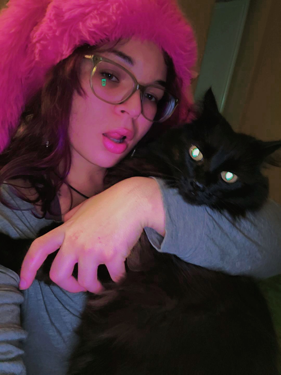 Young woman with glasses and pink fuzzy hat holding a black cat, both looking at the camera.
