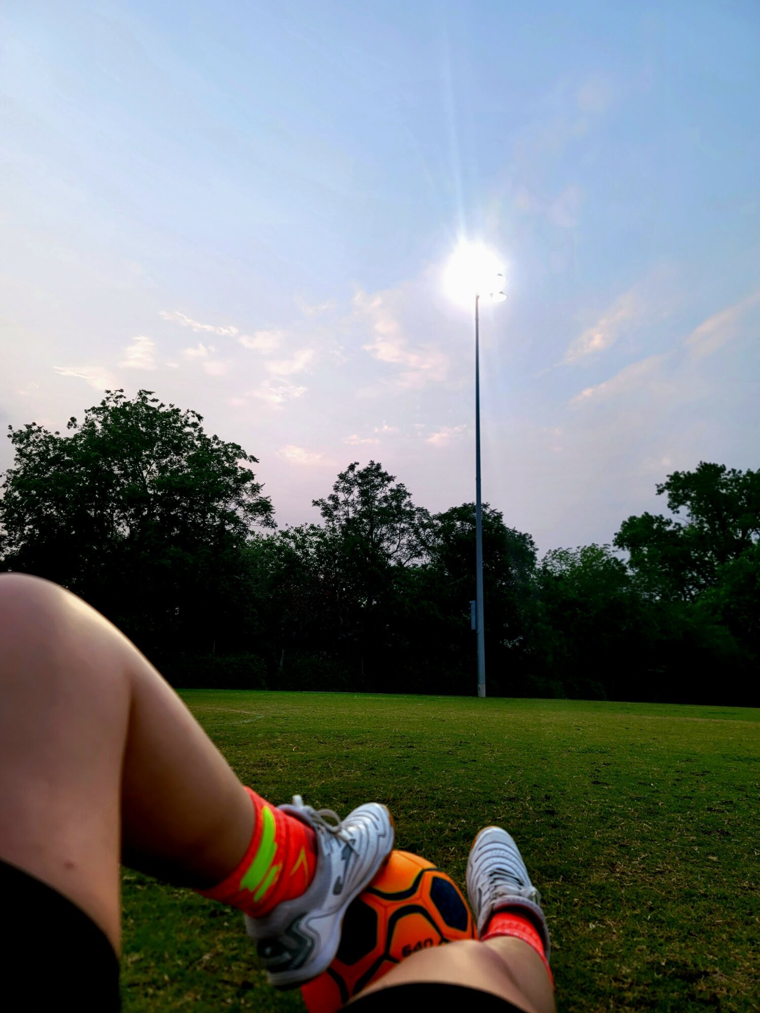 Person sitting on grass with legs extended, trees and a tall light pole in the background, sky with clouds.