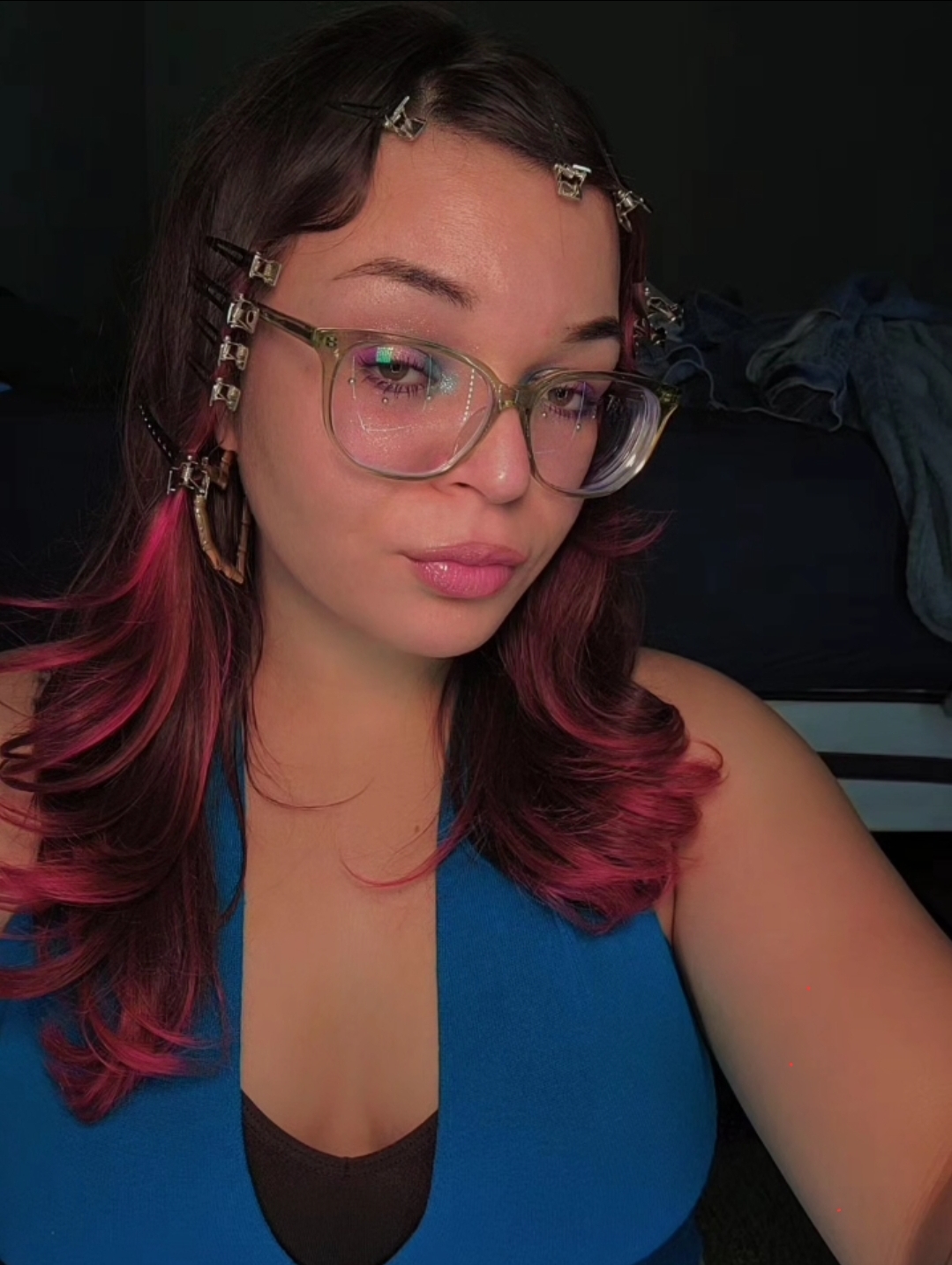 Young woman with glasses and pink-tinted hair, wearing a blue top, looking at the camera.