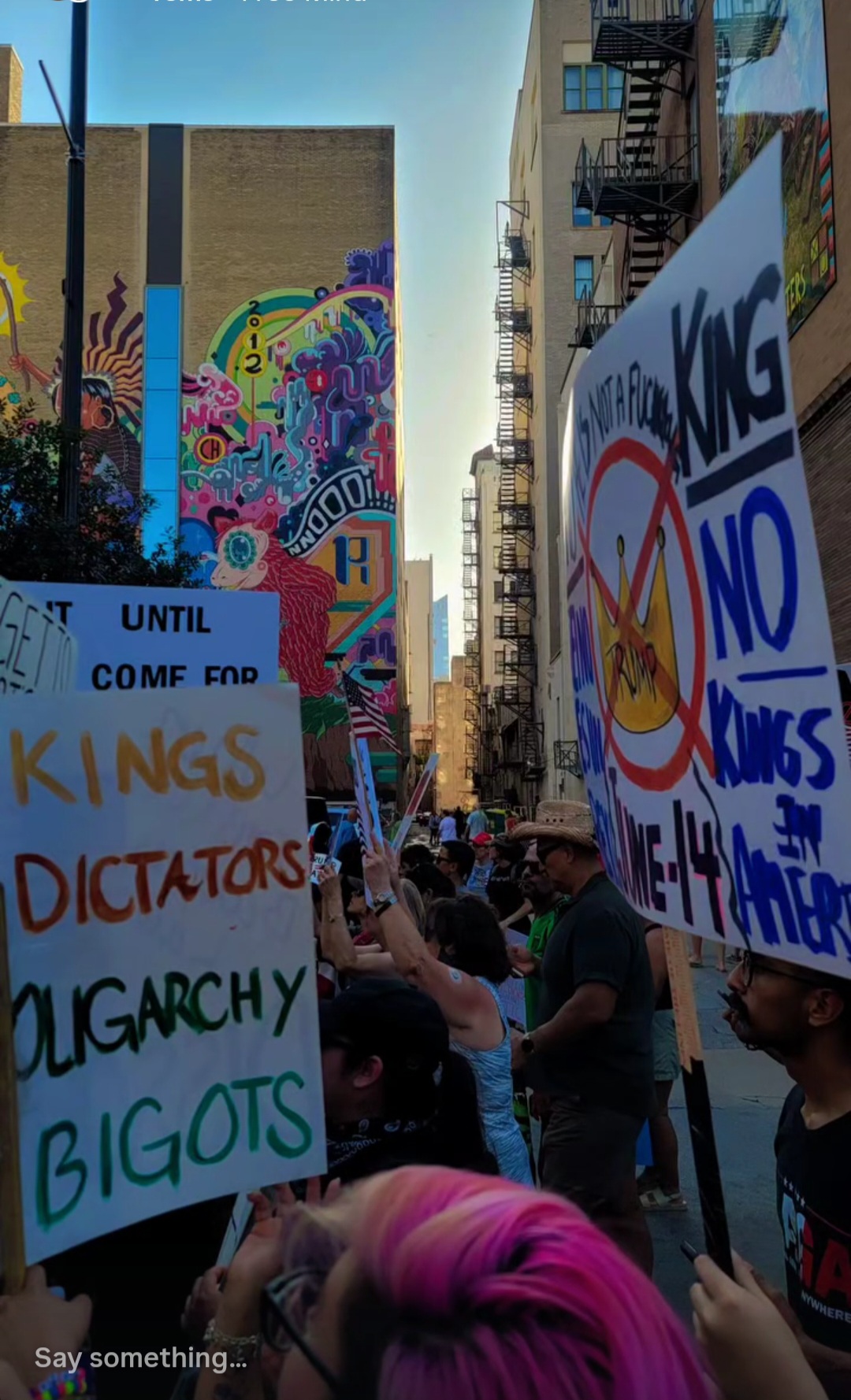 Protesters hold signs in a city alleyway with colorful graffiti on walls, tall buildings, and a clear sky above.