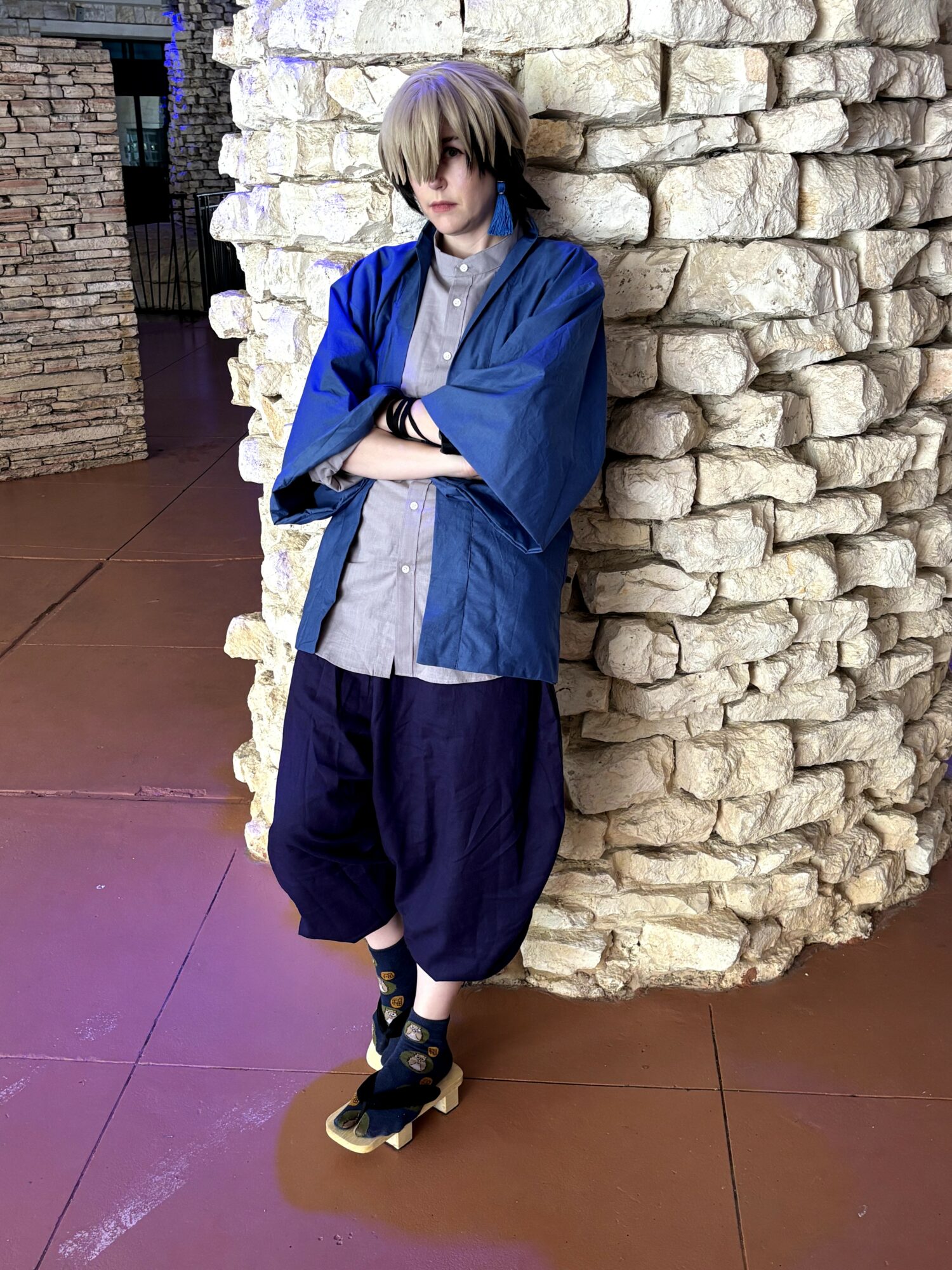 Person with short hair leaning against a stone wall, wearing a blue jacket, gray shirt, and black pants.