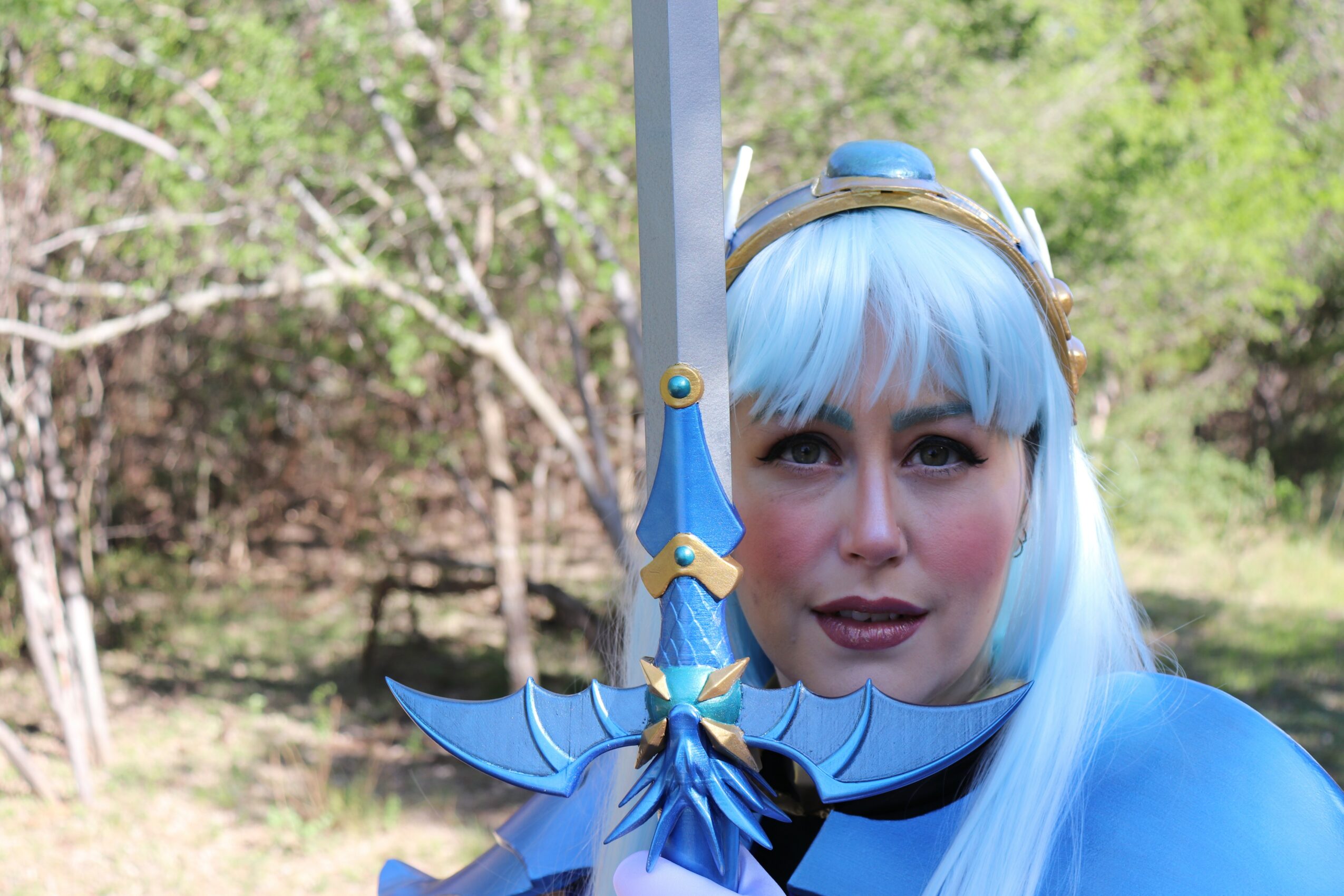 Person with blue hair and costume holding a sword outdoors, trees and greenery in background.
