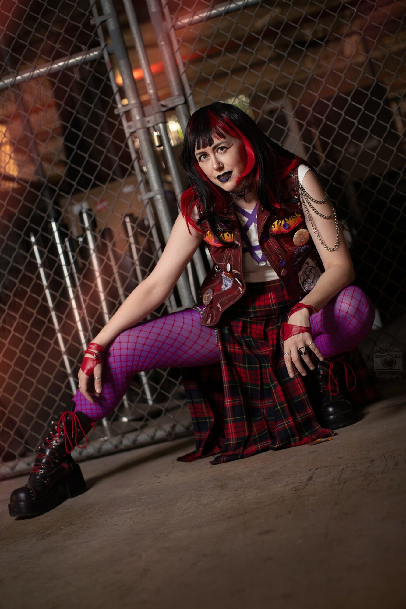 Young woman with dark hair and red highlights, wearing a plaid skirt, purple fishnet stockings, and a vest, sitting on the ground near a chain-link fence at night.