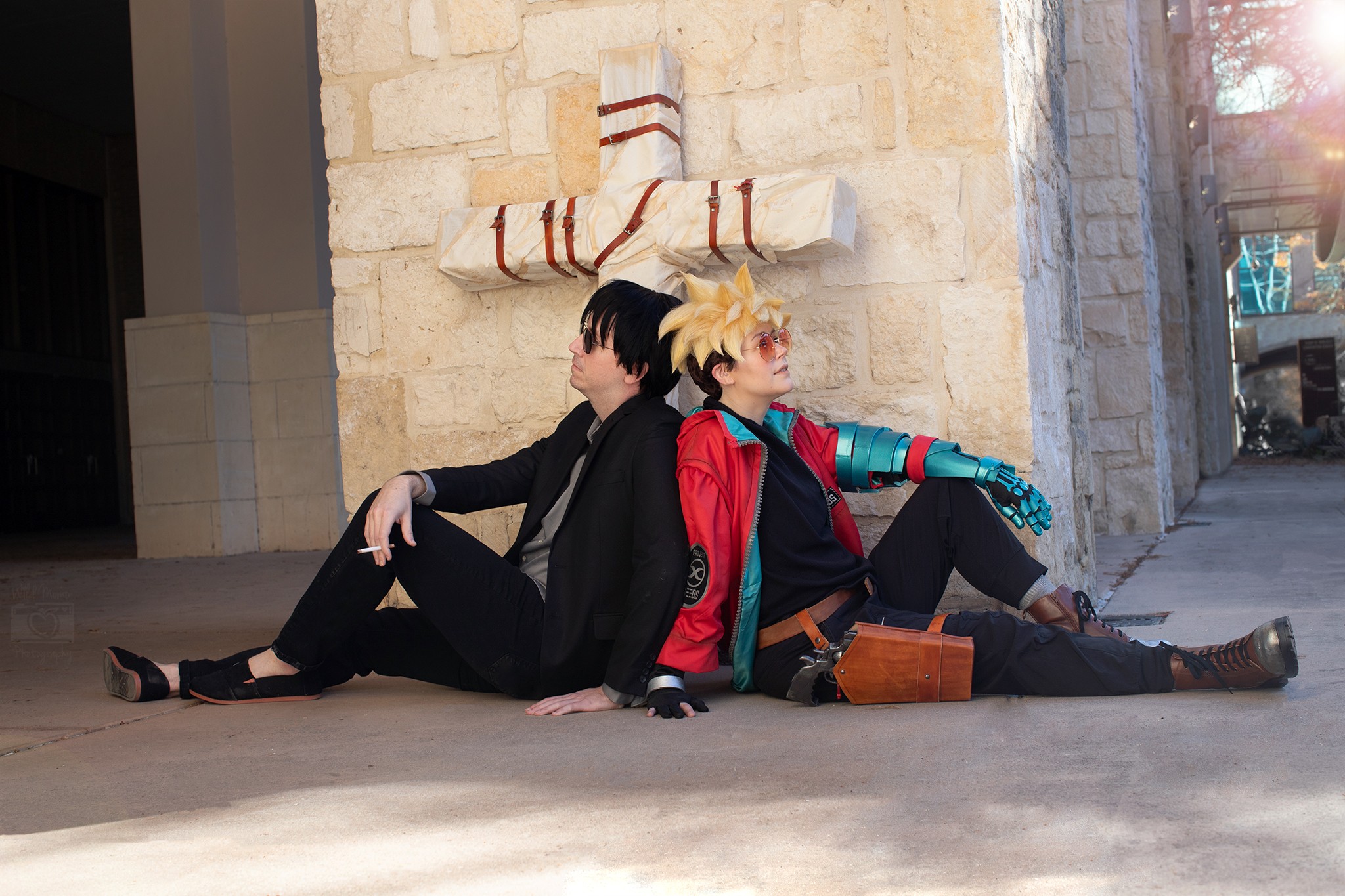 Two people sit back-to-back against a stone wall, one with black hair and the other with blonde hair, in cosplay costumes.