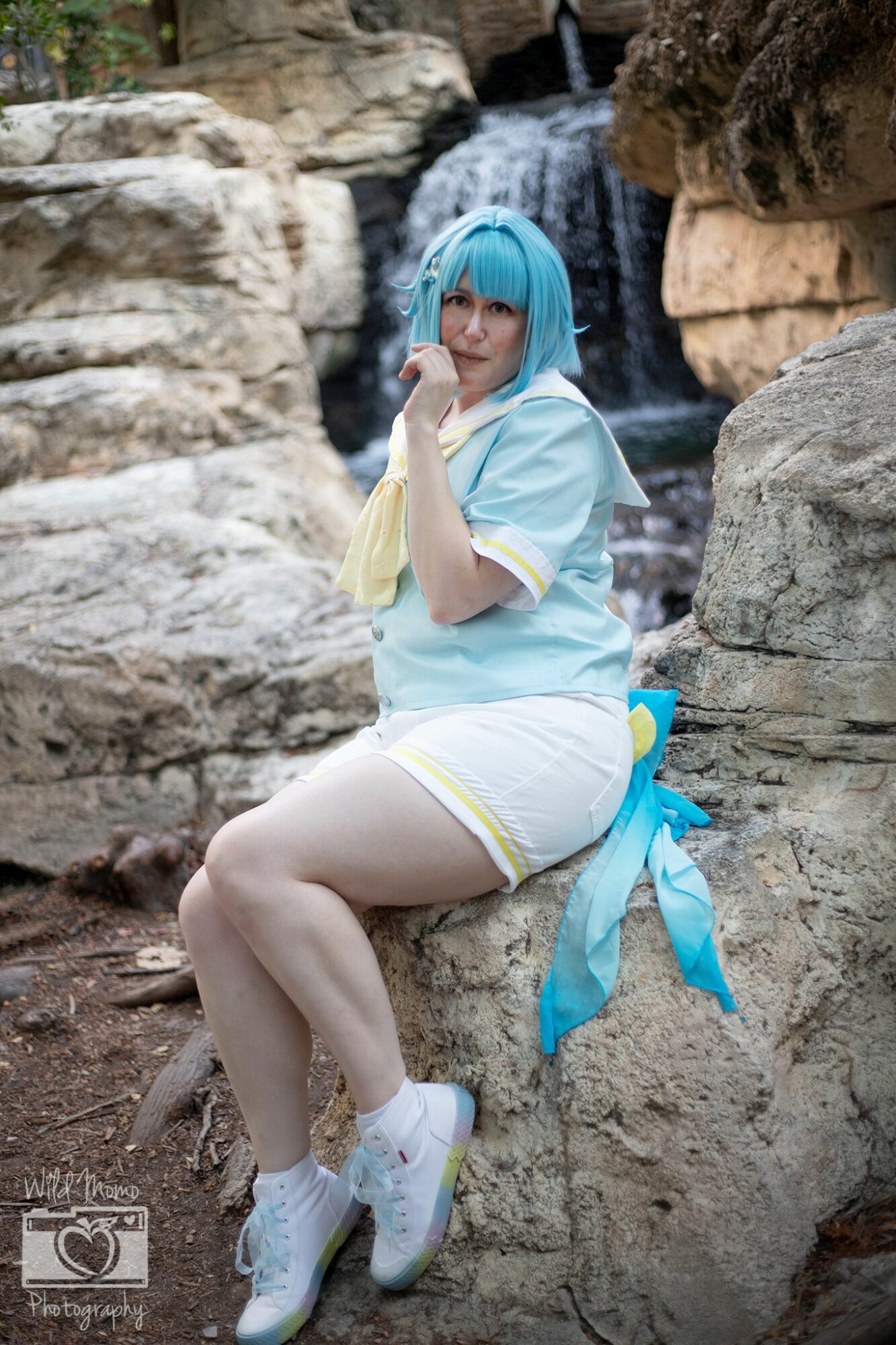 Person with blue wig and costume sitting on a rock in front of a waterfall, posing with finger near lips, outdoors.