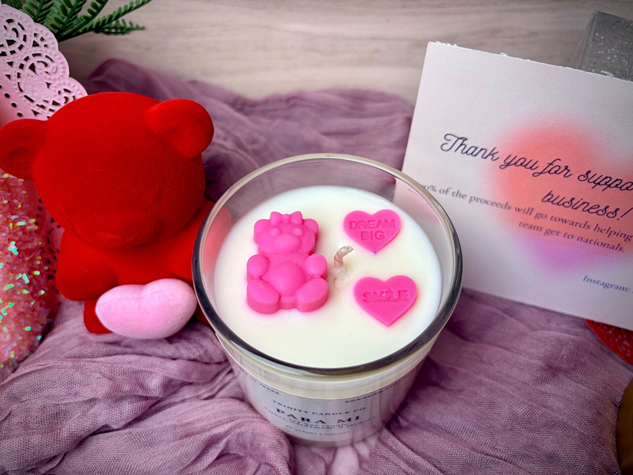 A candle with pink bear and heart-shaped decorations, a red plush bear, a pink plush heart, and a thank you card on purple fabric.
