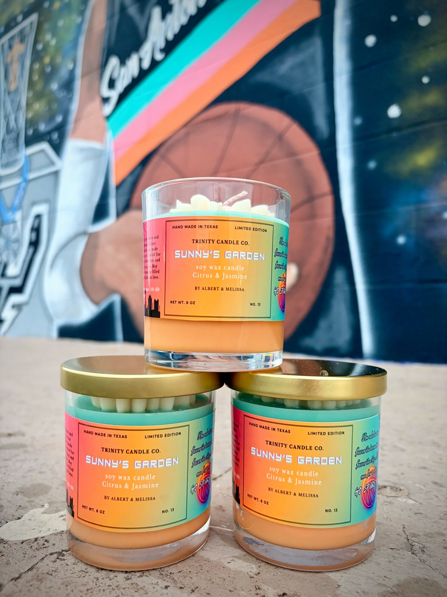 Three candles with colorful labels stacked on a surface, with a blurred background of a person and a mural.