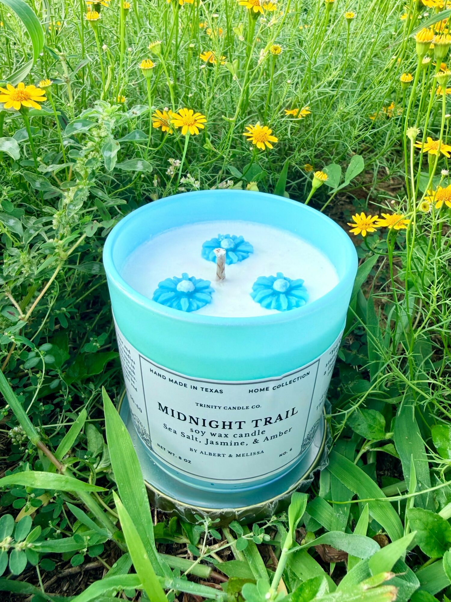 Blue candle with three flower-shaped decorations, labeled 'Midnight Trail,' placed among green grass and yellow flowers.
