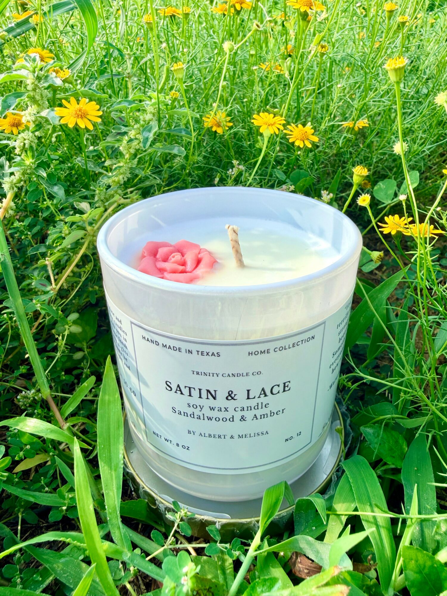 Satin and Lace soy wax candle with pink rose and beige wick, surrounded by green grass and yellow flowers outdoors.