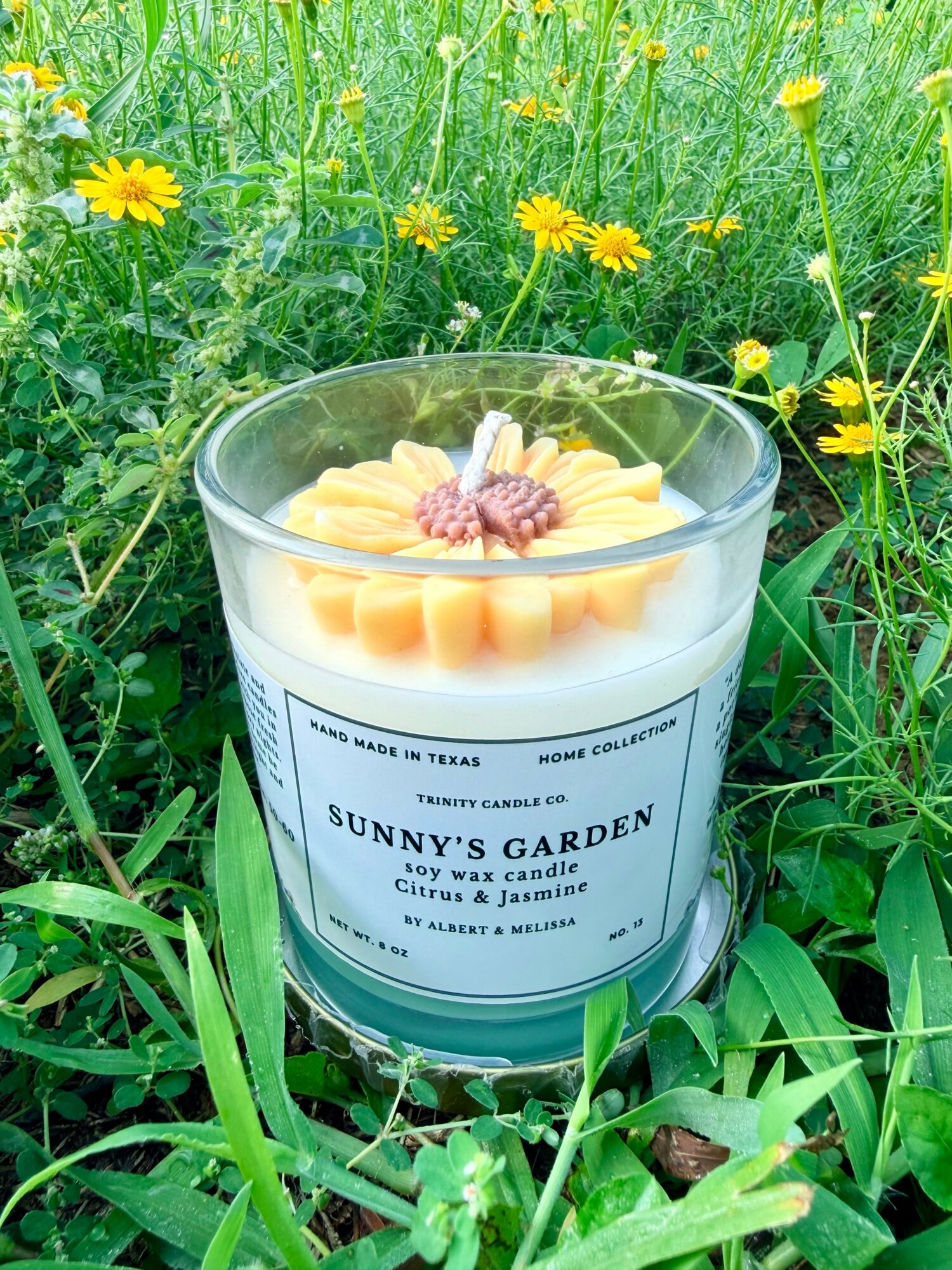 Scented candle with yellow flower-shaped wax, label reads Sunny's Garden, placed outdoors among green grass and yellow flowers.