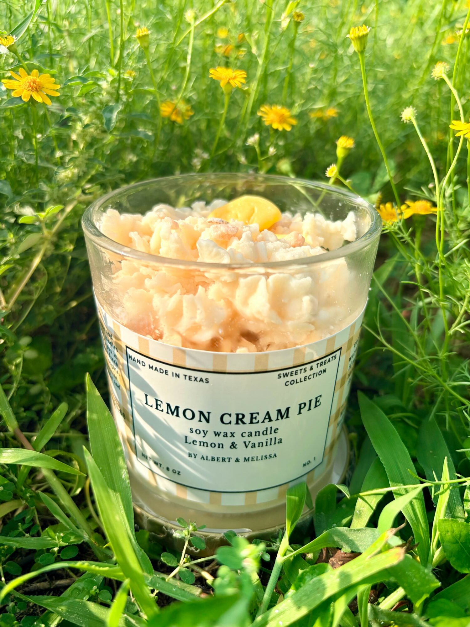 A candle labeled lemon cream pie in a glass jar, surrounded by green grass and yellow flowers outdoors.